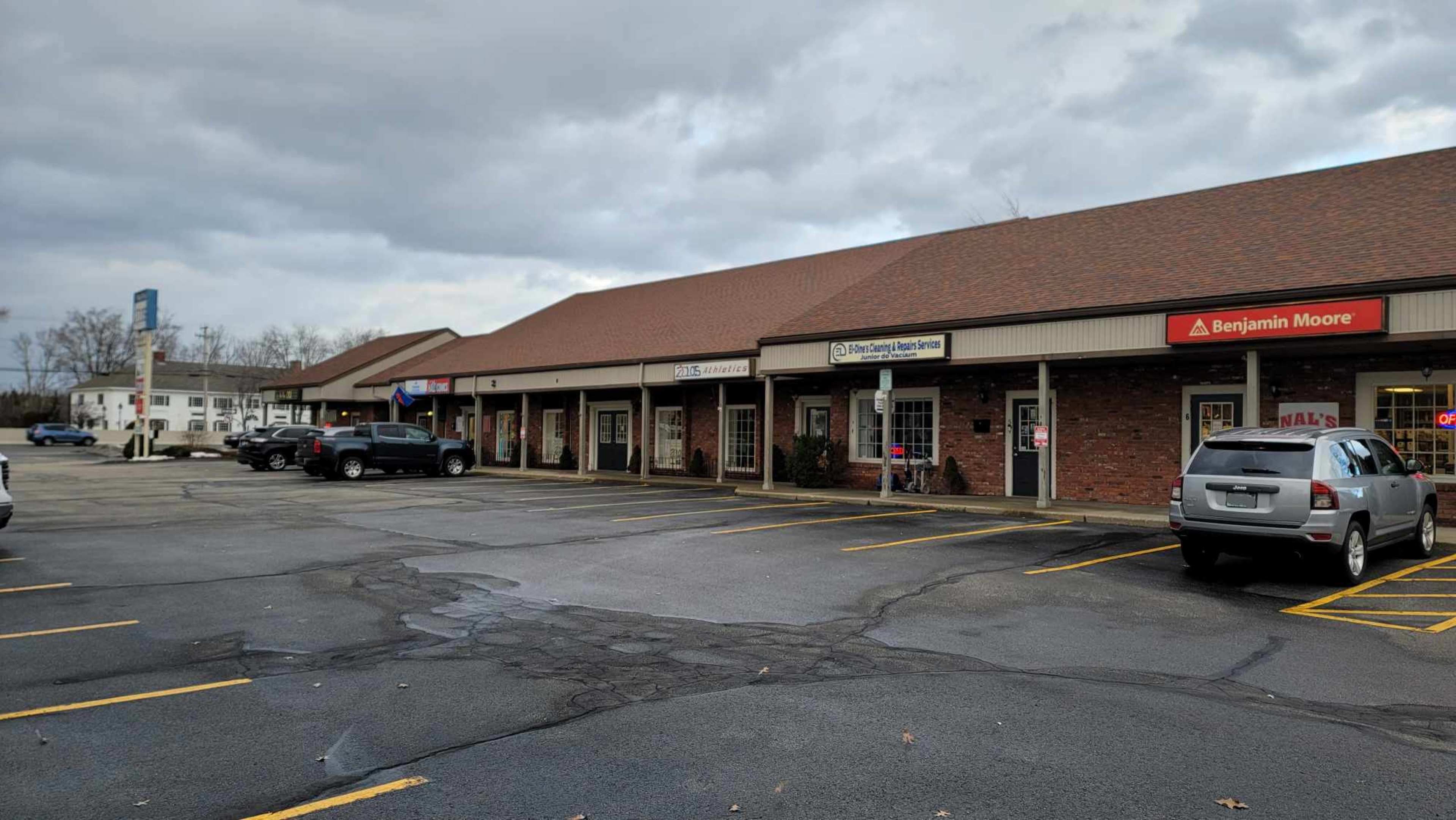 The scene shows a row of commercial storefronts in a parking lot with several cars parked in the spaces.
