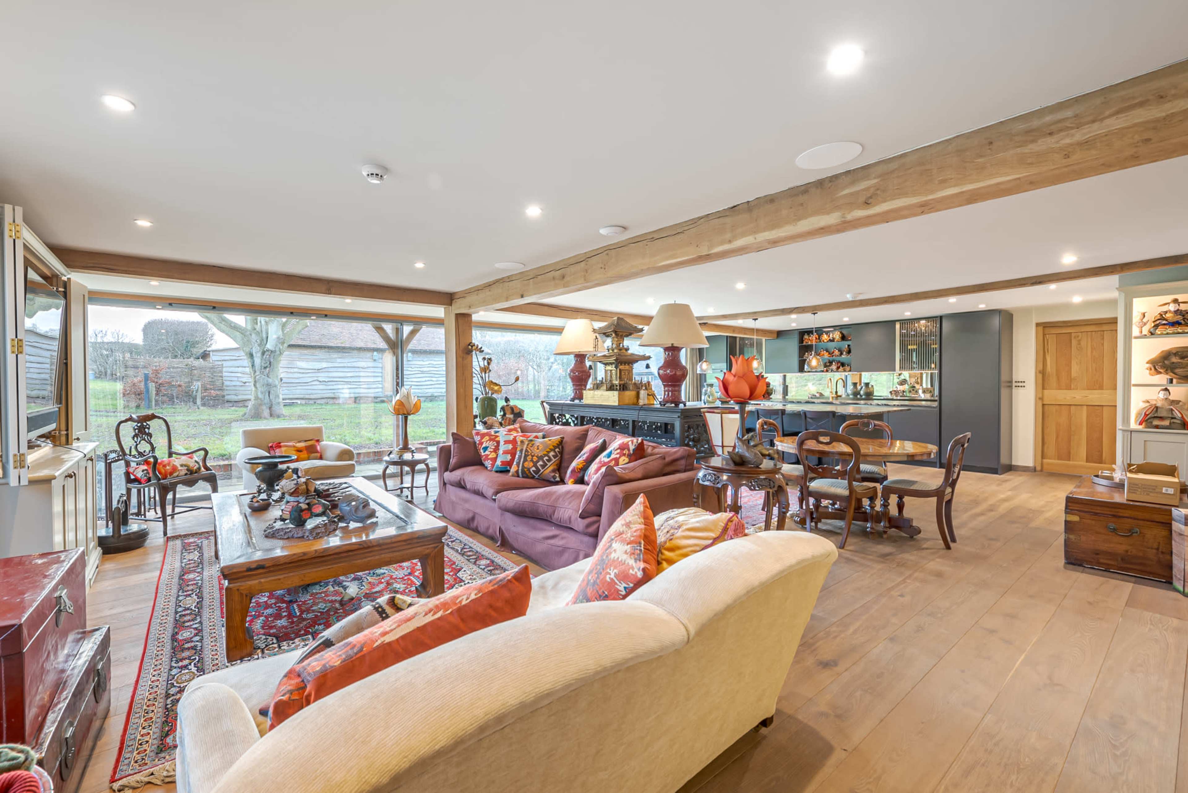 A spacious, open living area with a mix of seating arrangements, a dining table, and large windows that overlook a garden and a rustic barn.