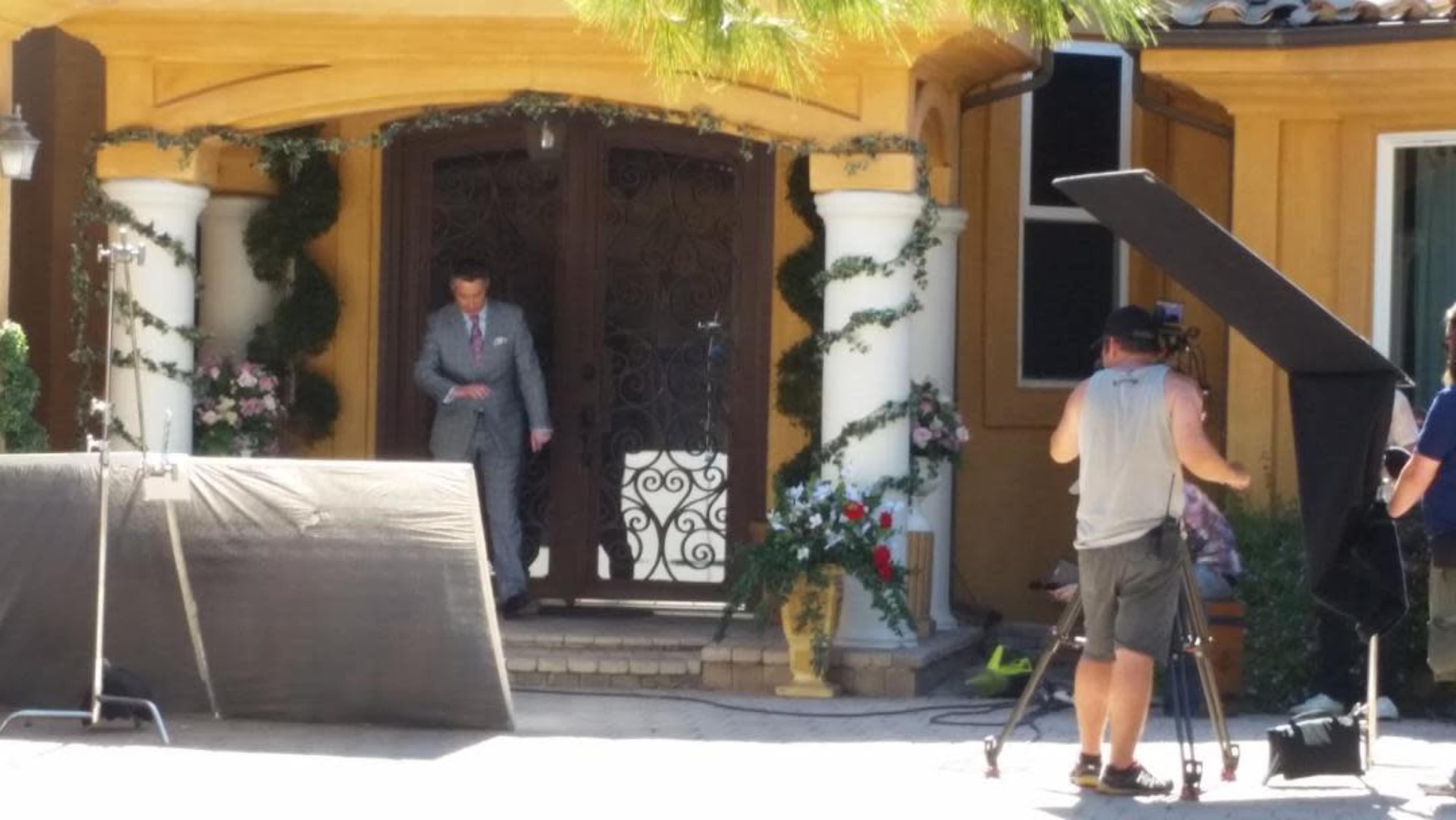 A man in a gray suit steps out of a grand entrance adorned with decorative plants while a film crew sets up equipment nearby.