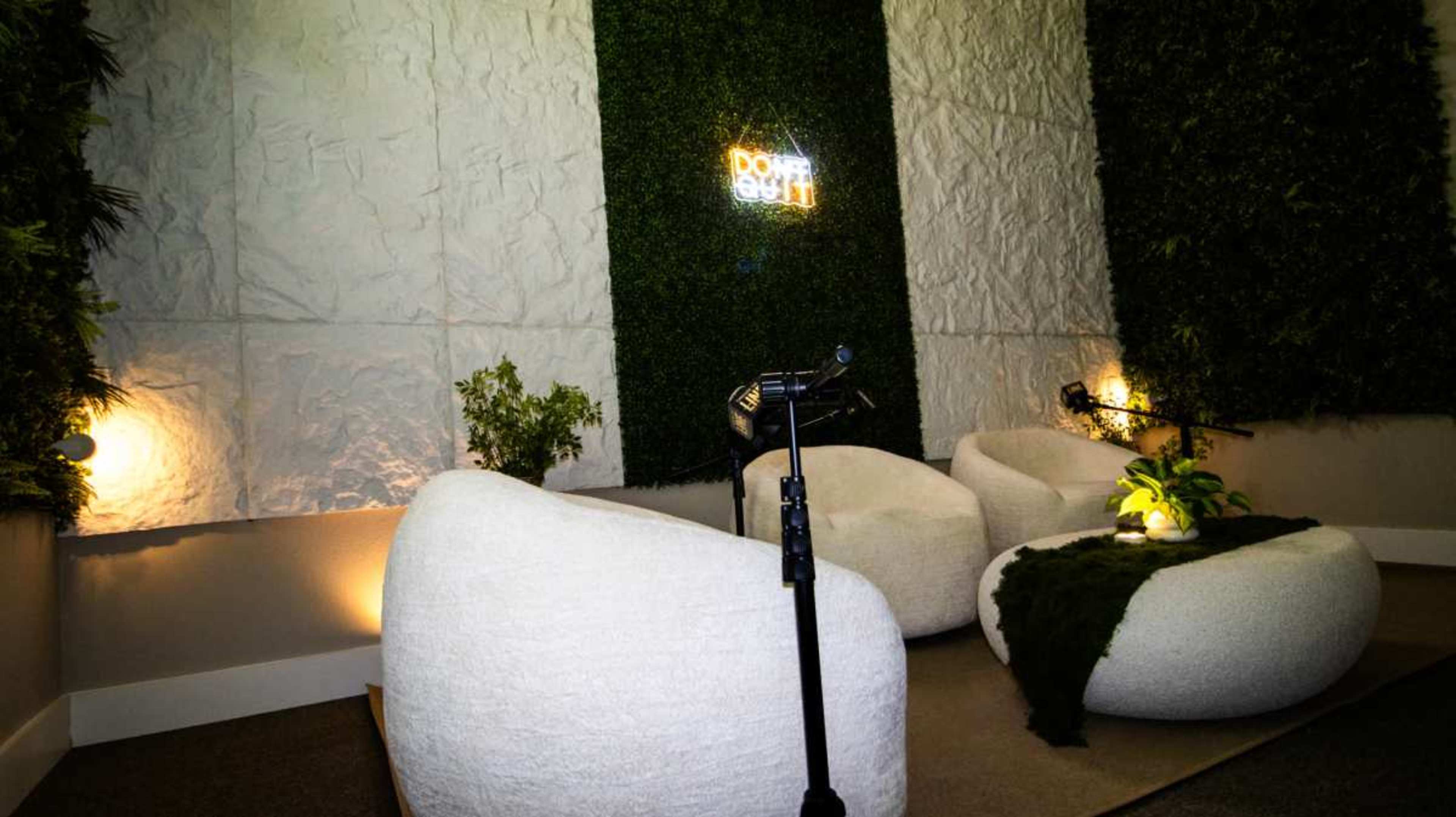 The image shows a modern, cozy seating area with two plush white chairs, a small plant, and decorative greenery on the walls, illuminated by soft lighting.