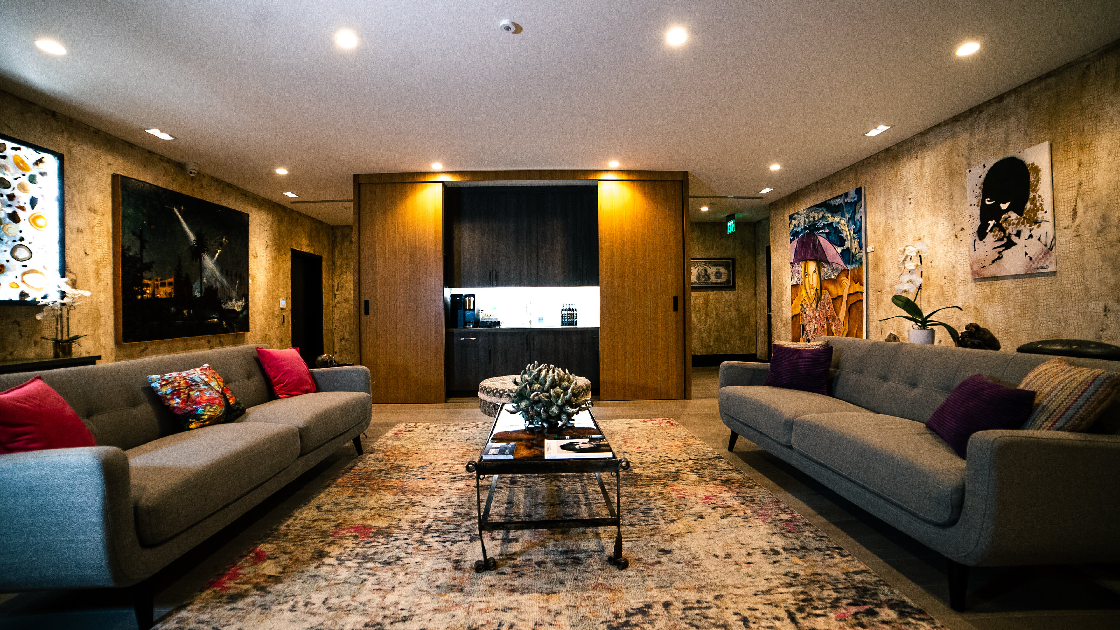 A well-lit living room with two sofas facing a coffee table, surrounded by artwork on the walls and a doorway leading to a kitchen area in the background.