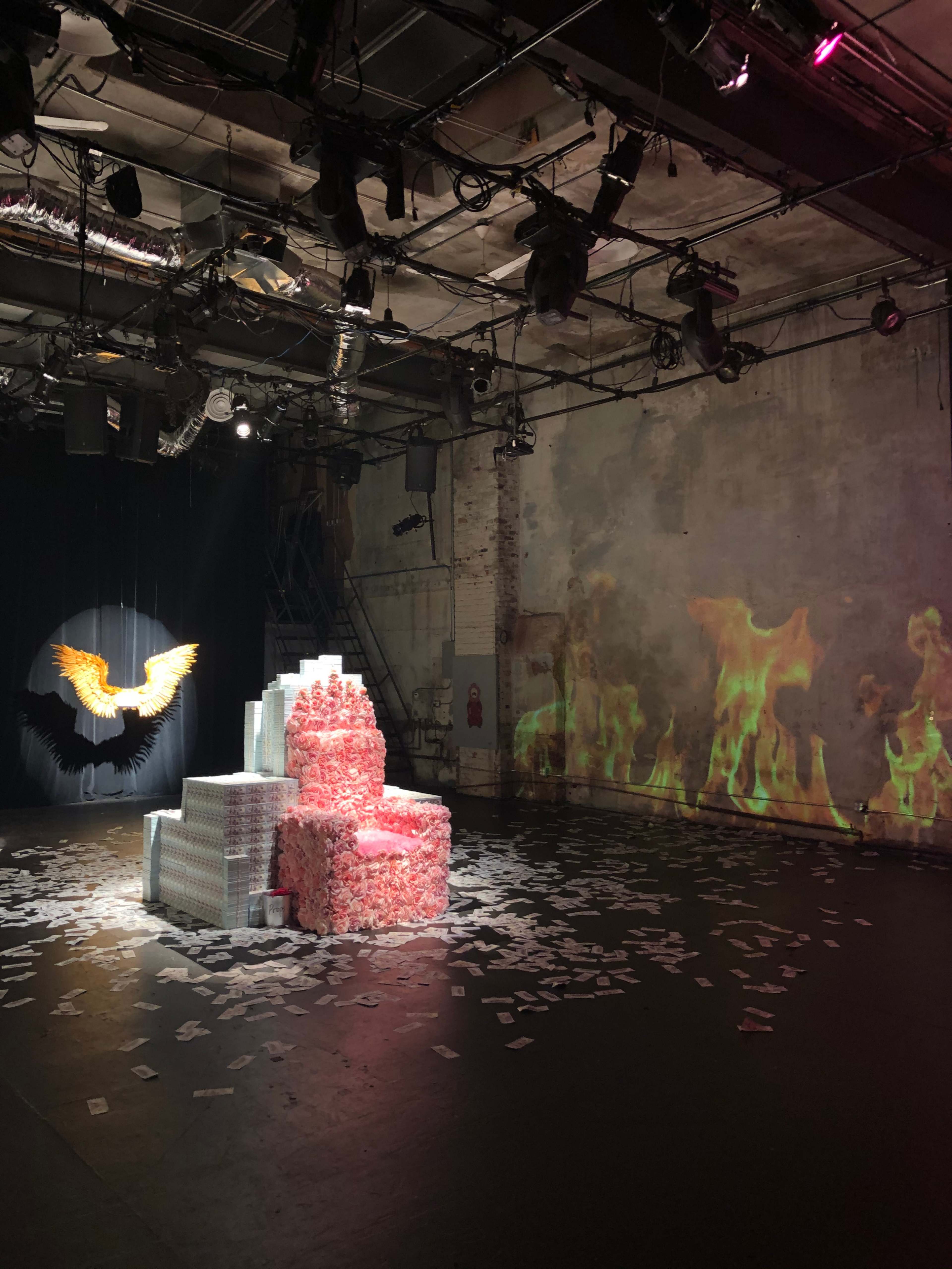 An art installation in a dimly lit space featuring a stack of decorated boxes, a backdrop of projected flames, and scattered papers on the floor.