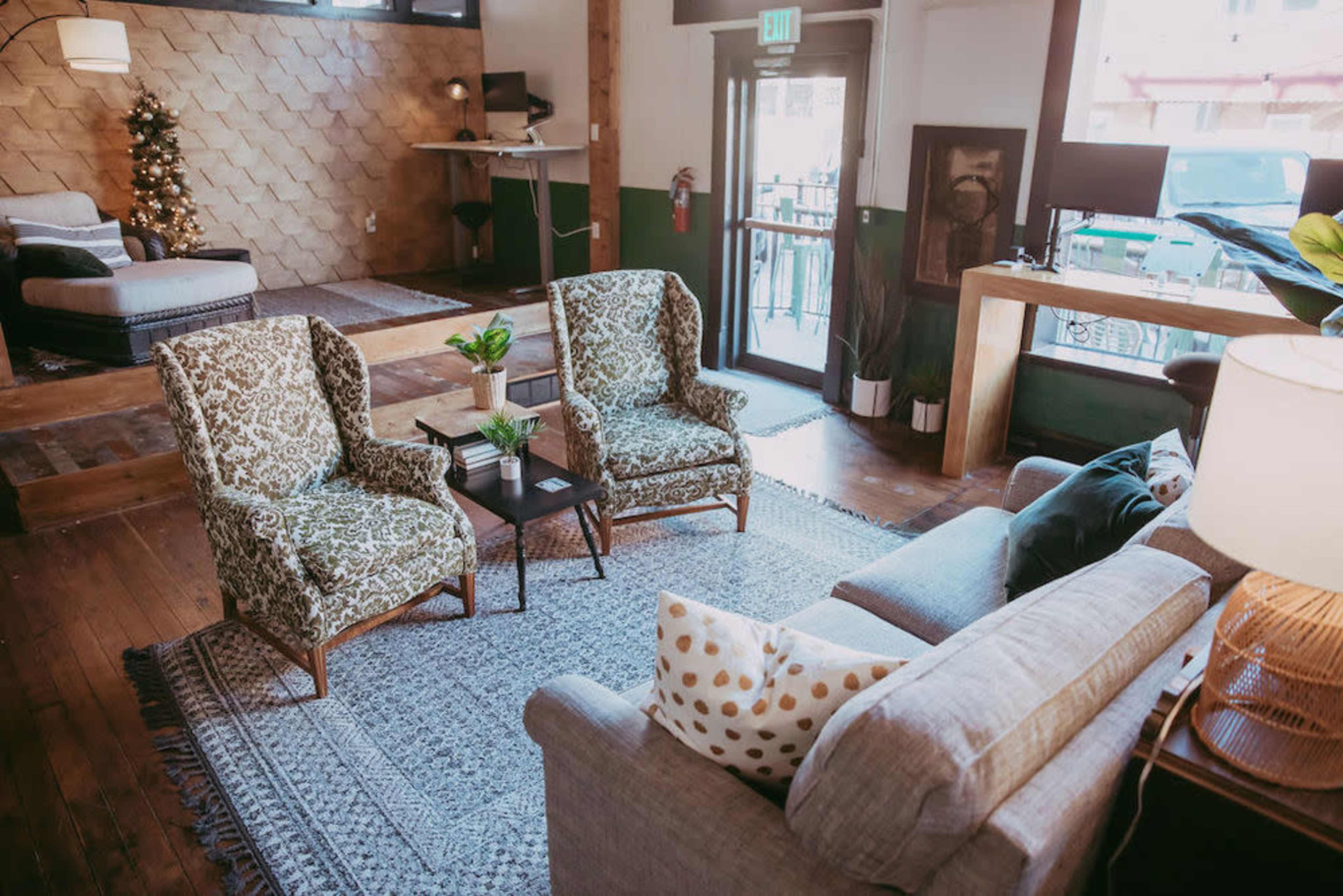 The image shows a cozy lounge area with two patterned armchairs and a sofa, surrounded by wooden furniture and a small table, in a well-lit space featuring a Christmas tree.