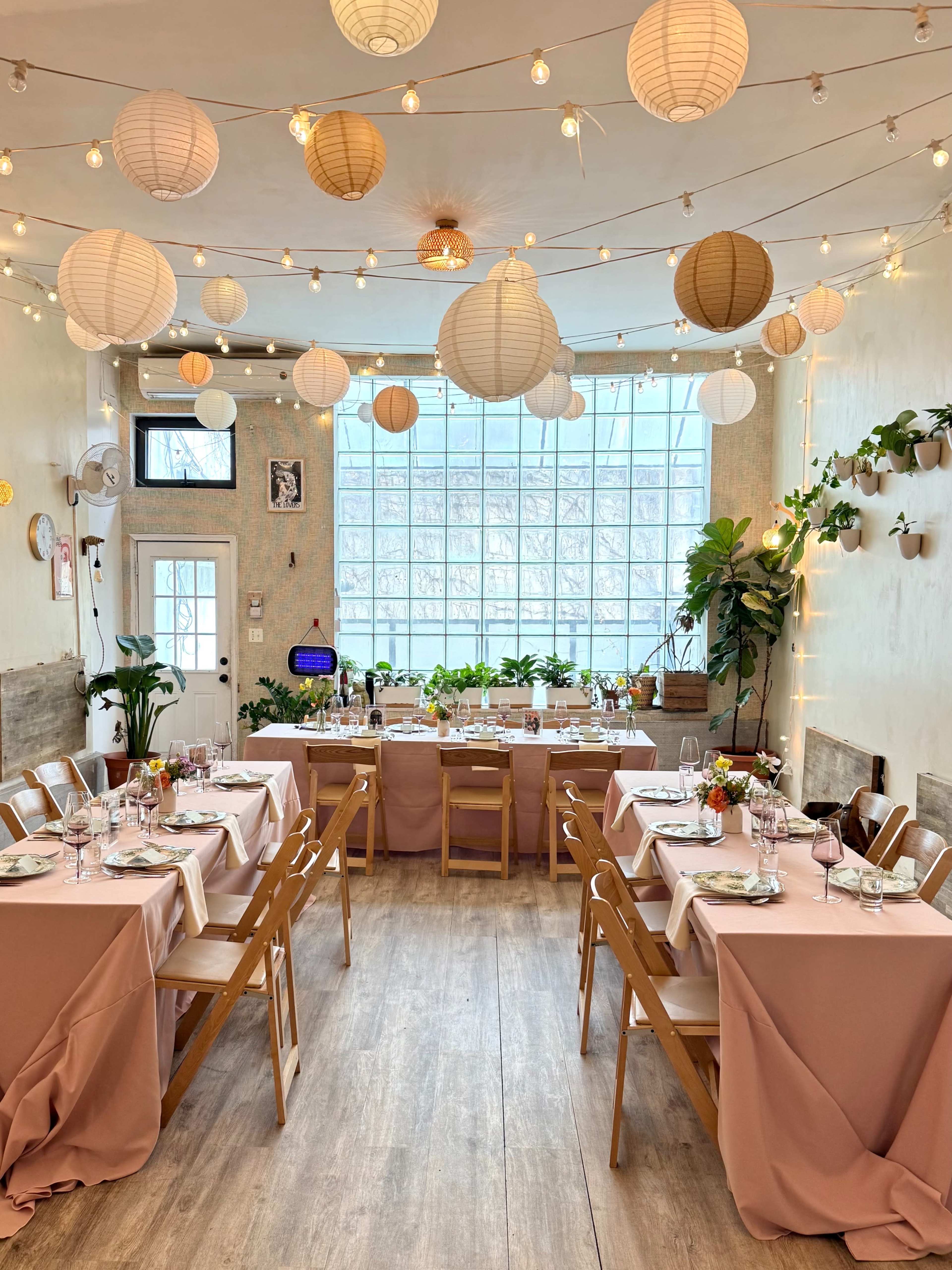 The image shows a decorated dining area with tables set for a meal, featuring pink tablecloths, plants, and hanging lanterns.
