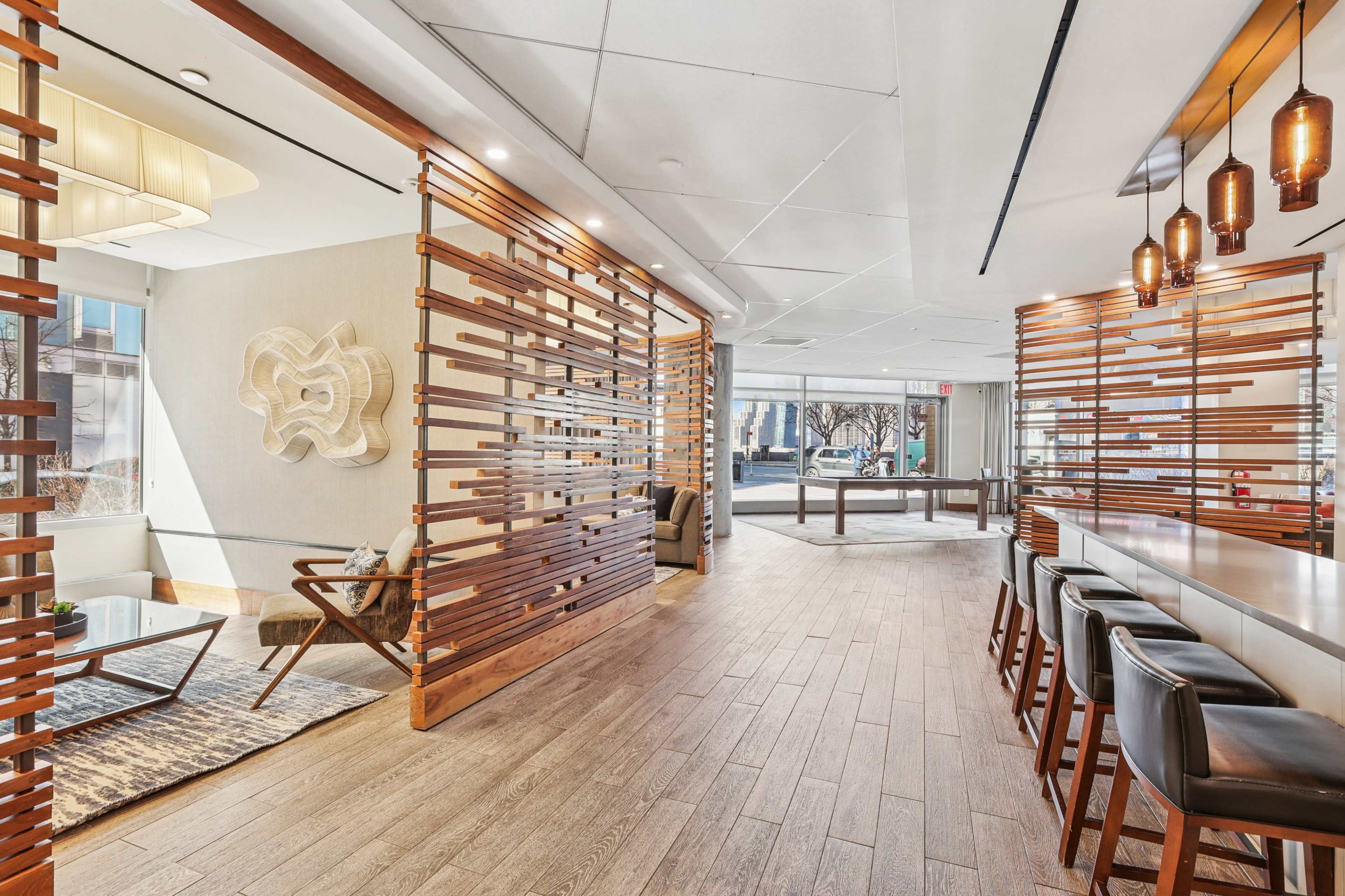 The image depicts a modern interior space with wooden slat partitions, a long table with high chairs, and a seating area featuring contemporary furniture.