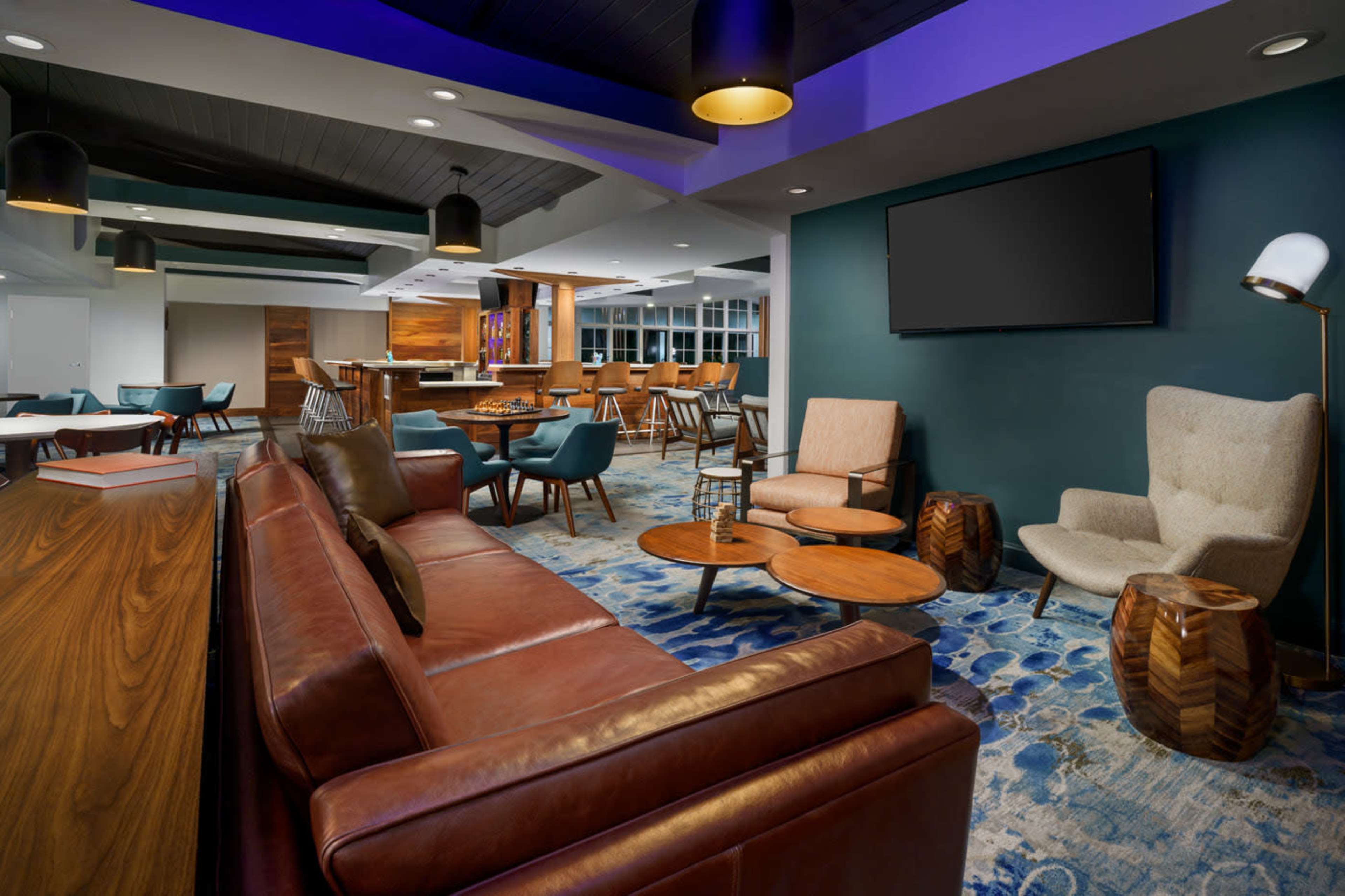 The image shows a modern lounge area with leather and fabric seating, wooden tables, and a bar in the background.