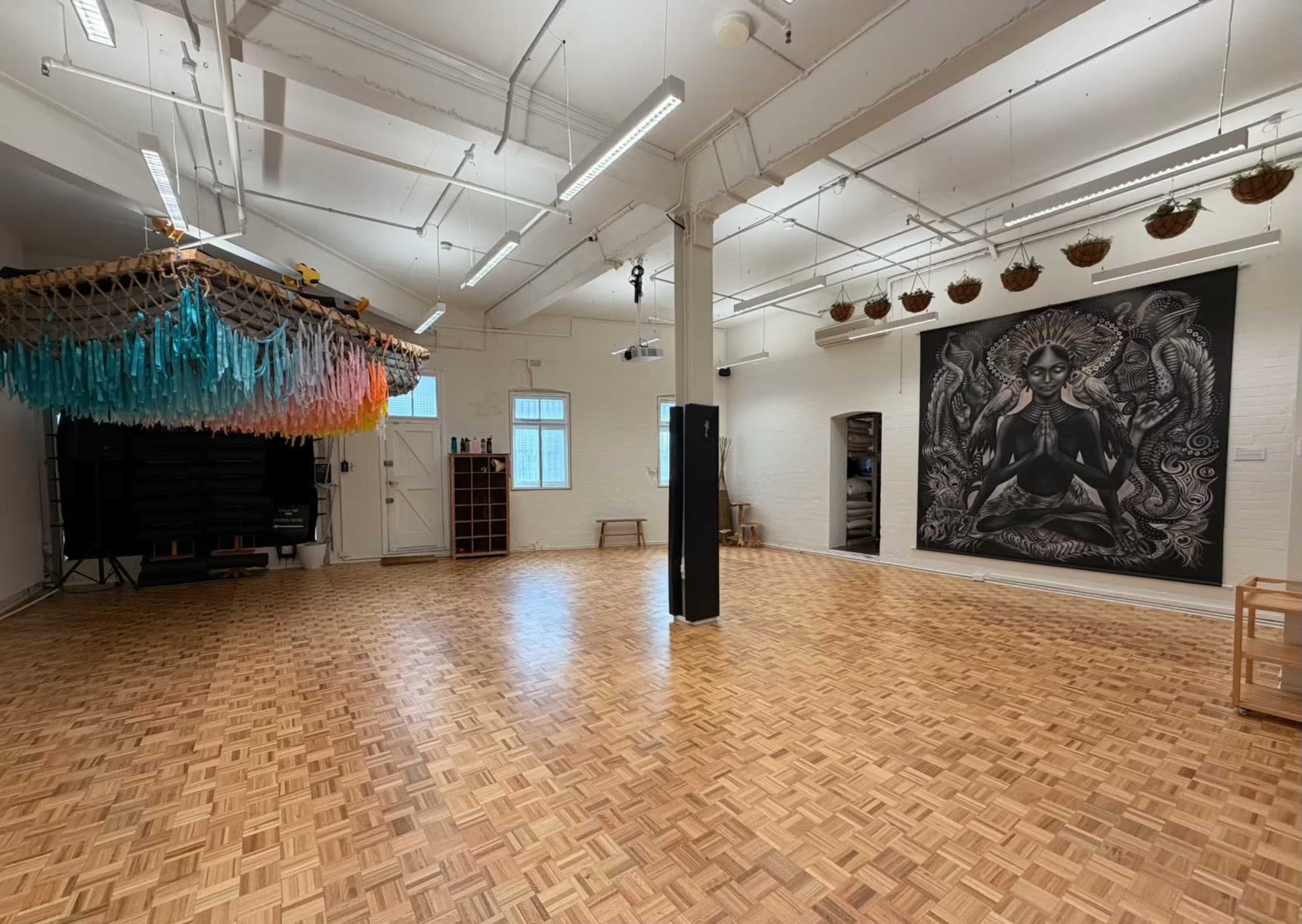 The image shows a spacious room with wooden flooring, featuring a large mural on one wall and colorful hanging decor overhead.