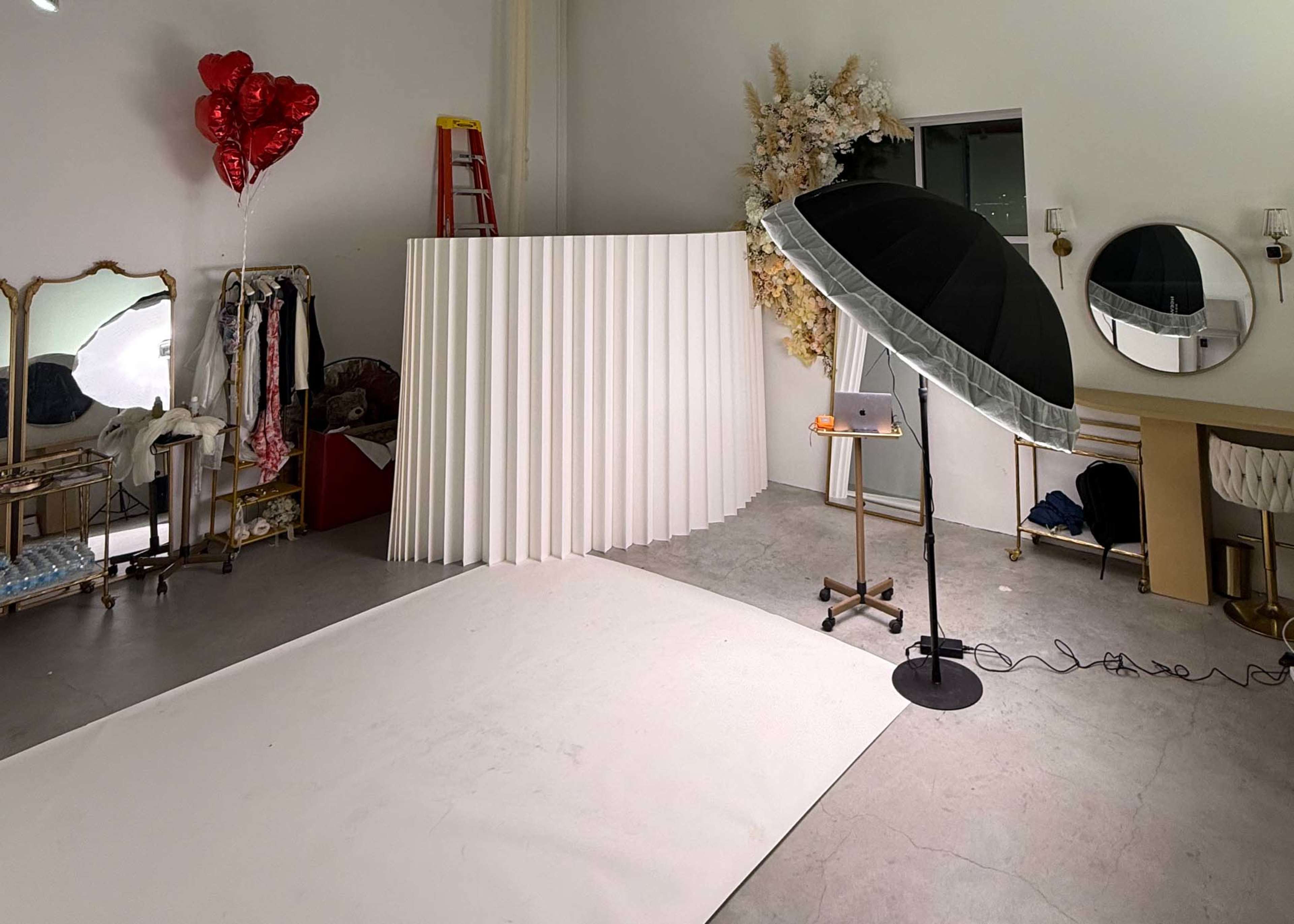 The image shows a spacious studio setup with a white backdrop, photography equipment, a ladder, and decorative elements like balloons and plants.