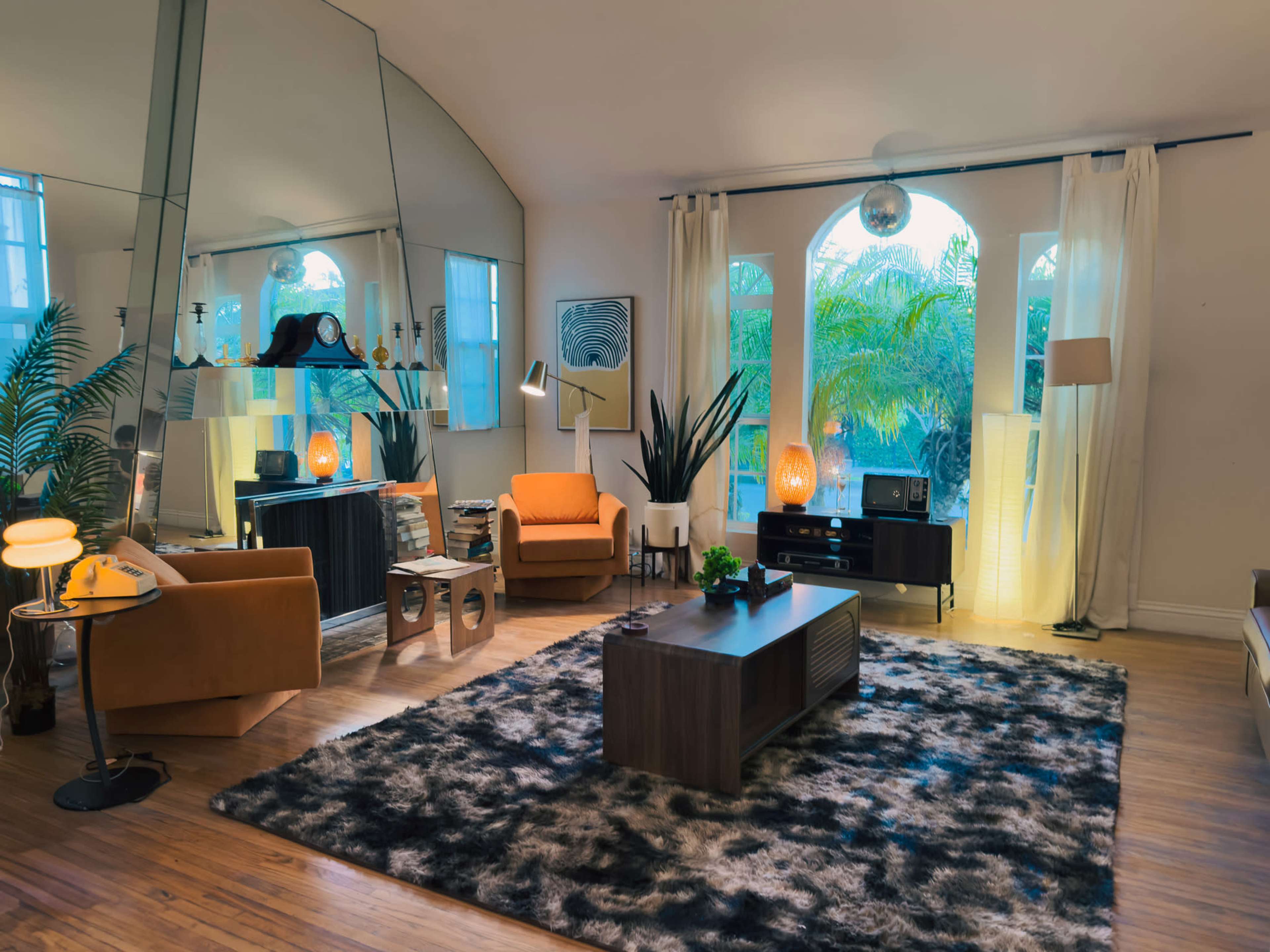The image depicts a stylish living room featuring a large mirror, modern furniture, and tropical plants under soft lighting.
