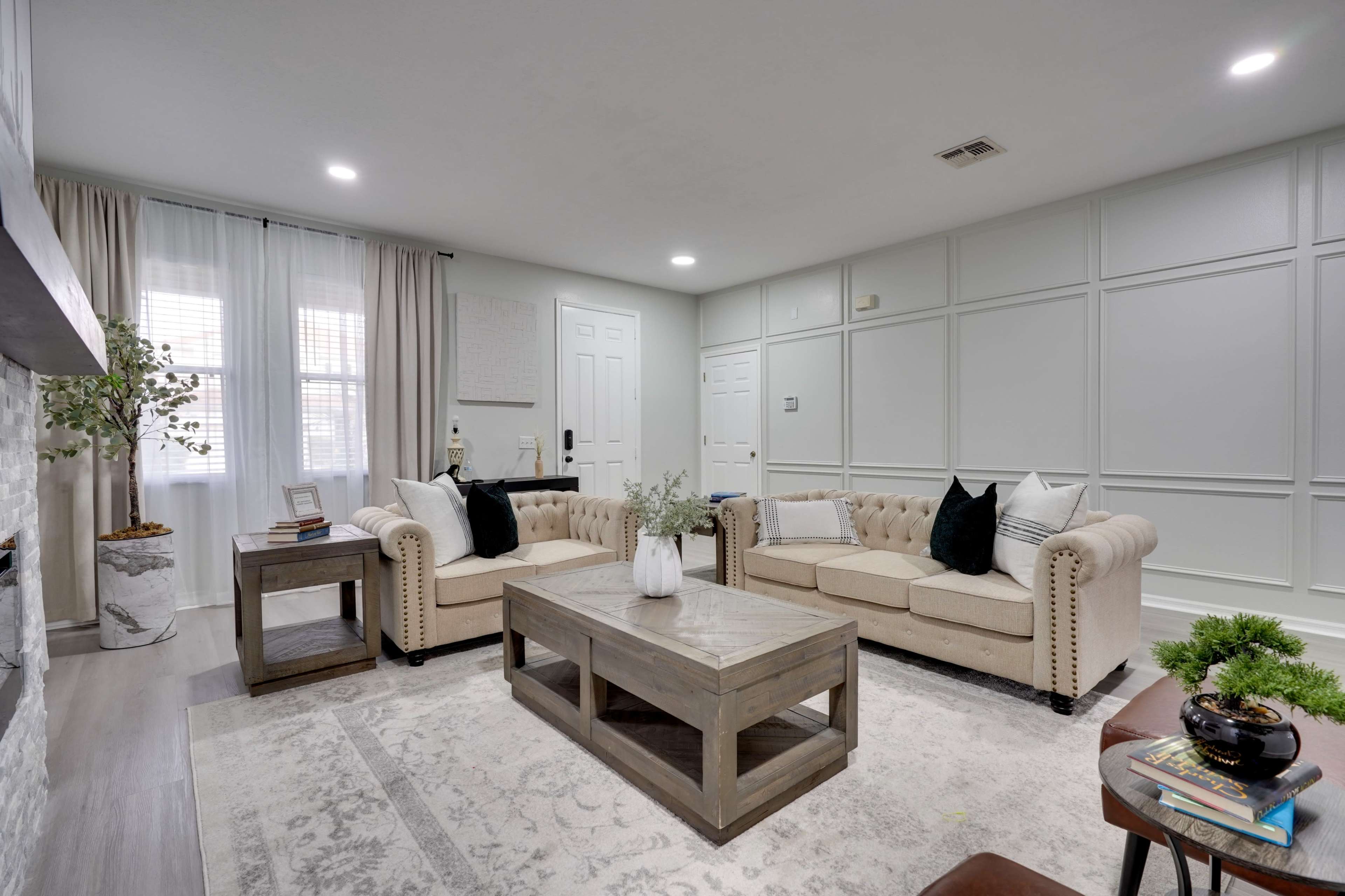 A living room features two beige tufted sofas facing a wooden coffee table, with a patterned rug on the floor and wall paneling in the background.