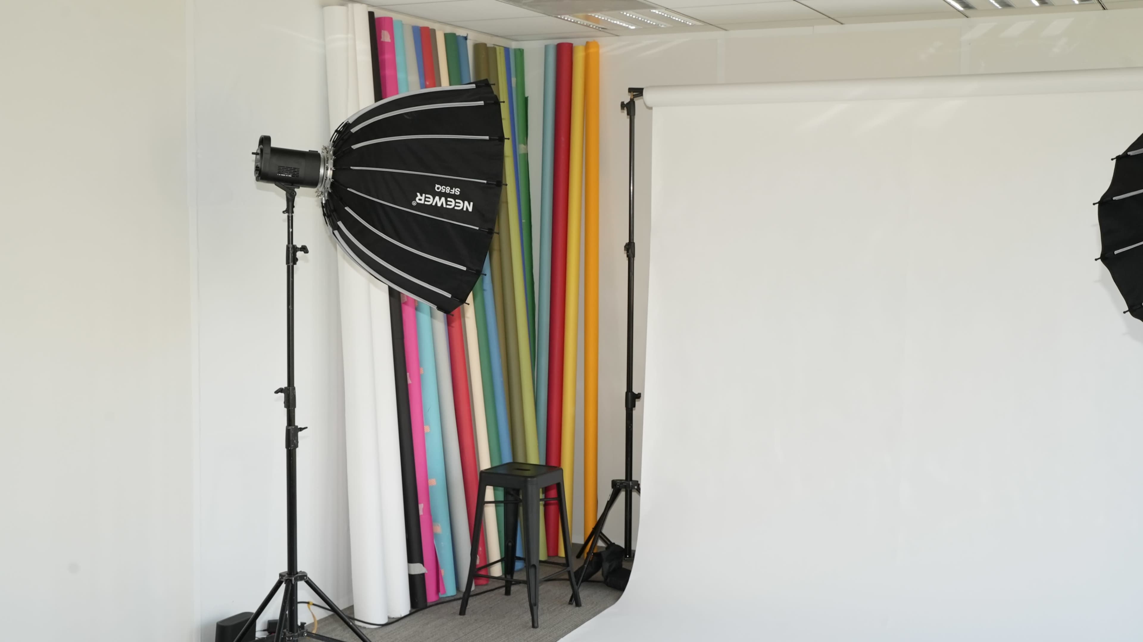 A photography studio is equipped with a lighting setup and a blank white backdrop, alongside a colorful roll of backdrop paper.