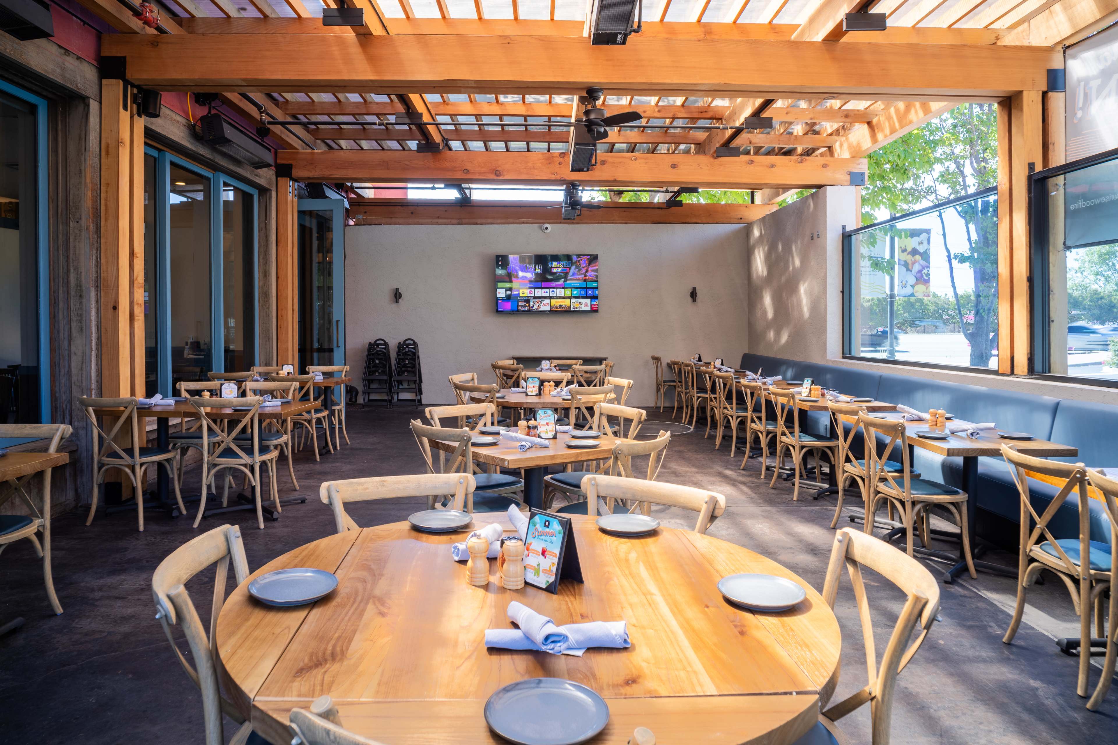 The image shows a spacious restaurant with wooden tables and chairs, large windows, and a TV mounted on the wall, designed for indoor dining.