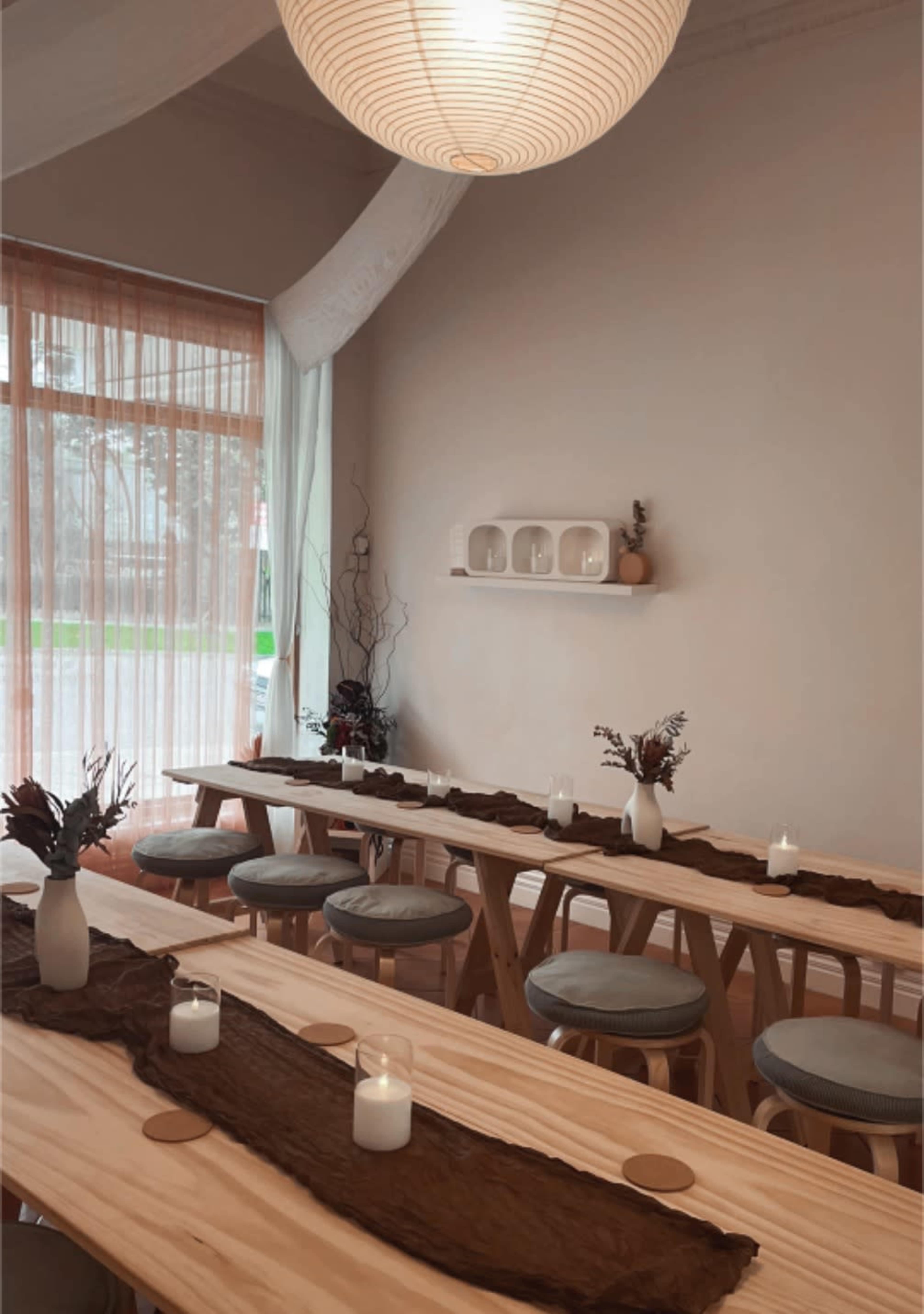 The image shows a serene dining space with wooden tables, minimalist decor, and soft lighting, featuring candles and simple floral arrangements.