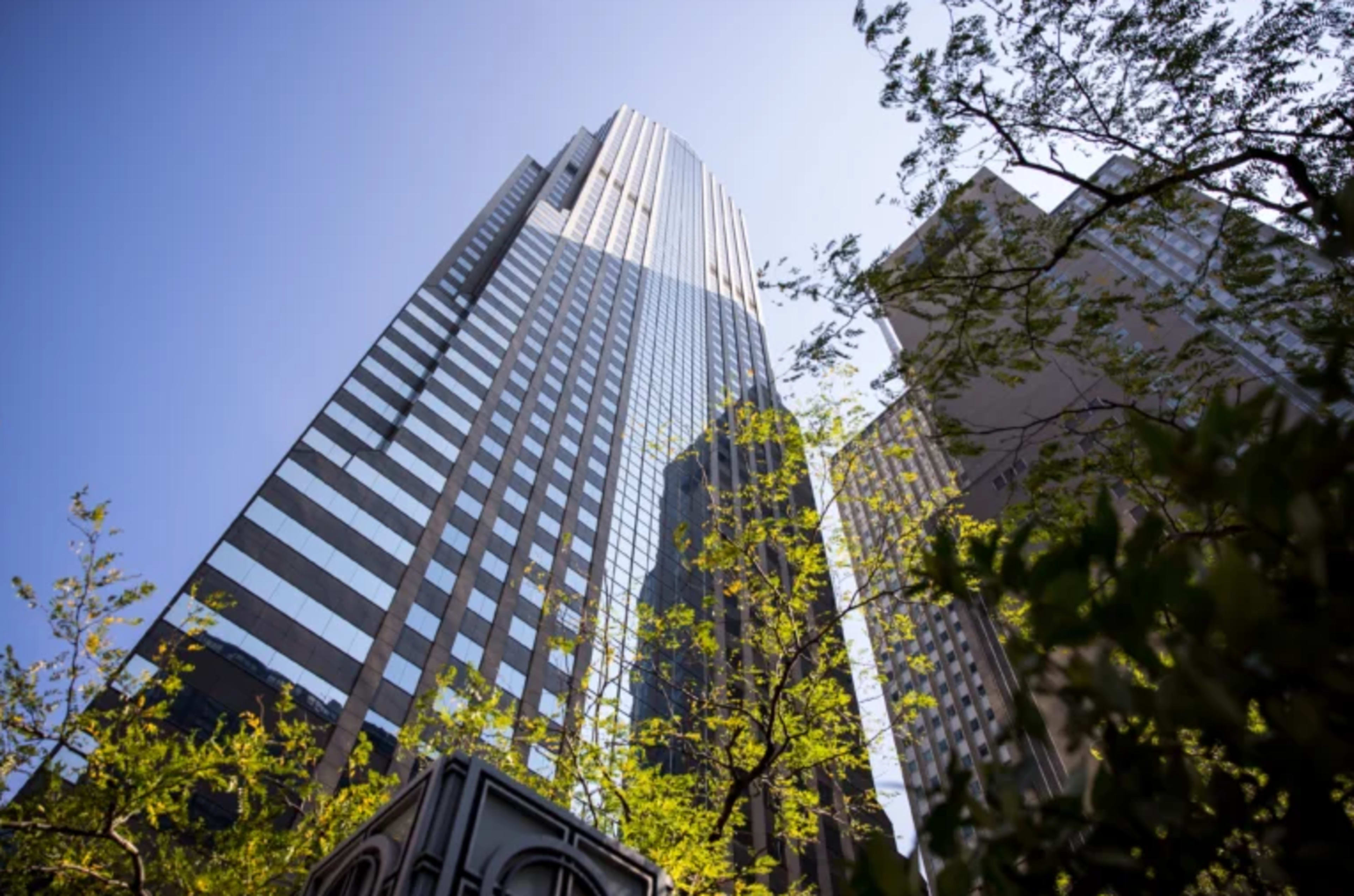 The image shows a tall skyscraper with a reflective facade, surrounded by trees and other buildings under a clear blue sky.
