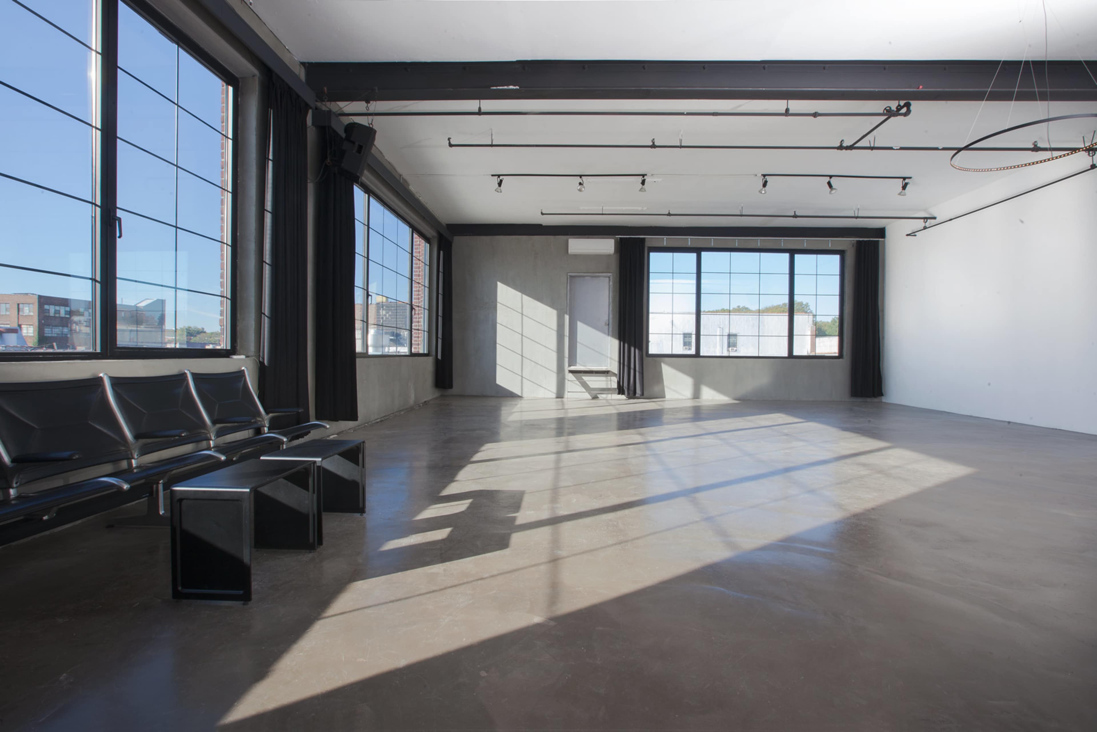 An empty industrial-style room with large windows, polished concrete flooring, and a few black chairs against one wall.