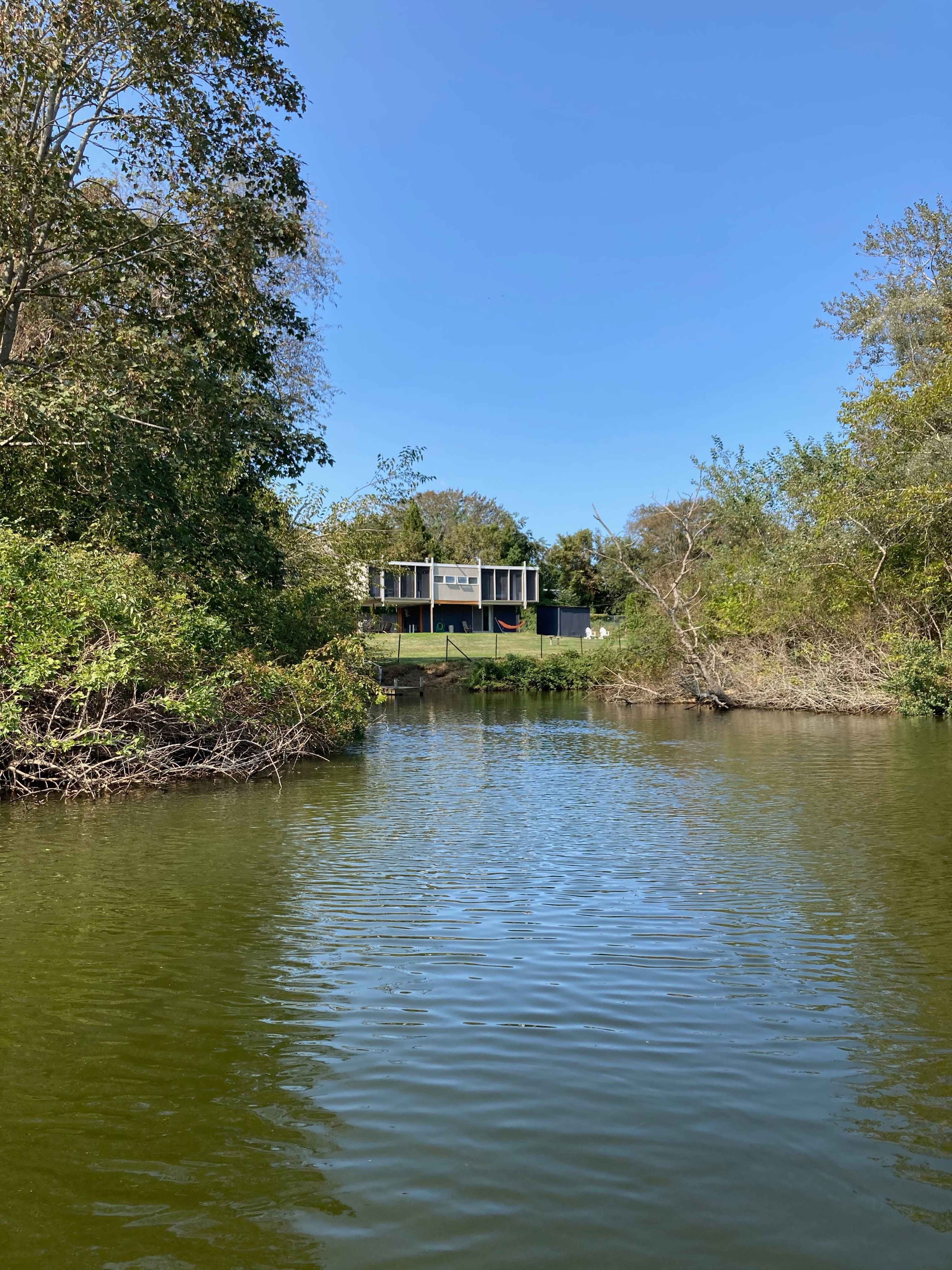 A modern house is situated on a grassy bank beside a calm river, surrounded by trees and shrubs.