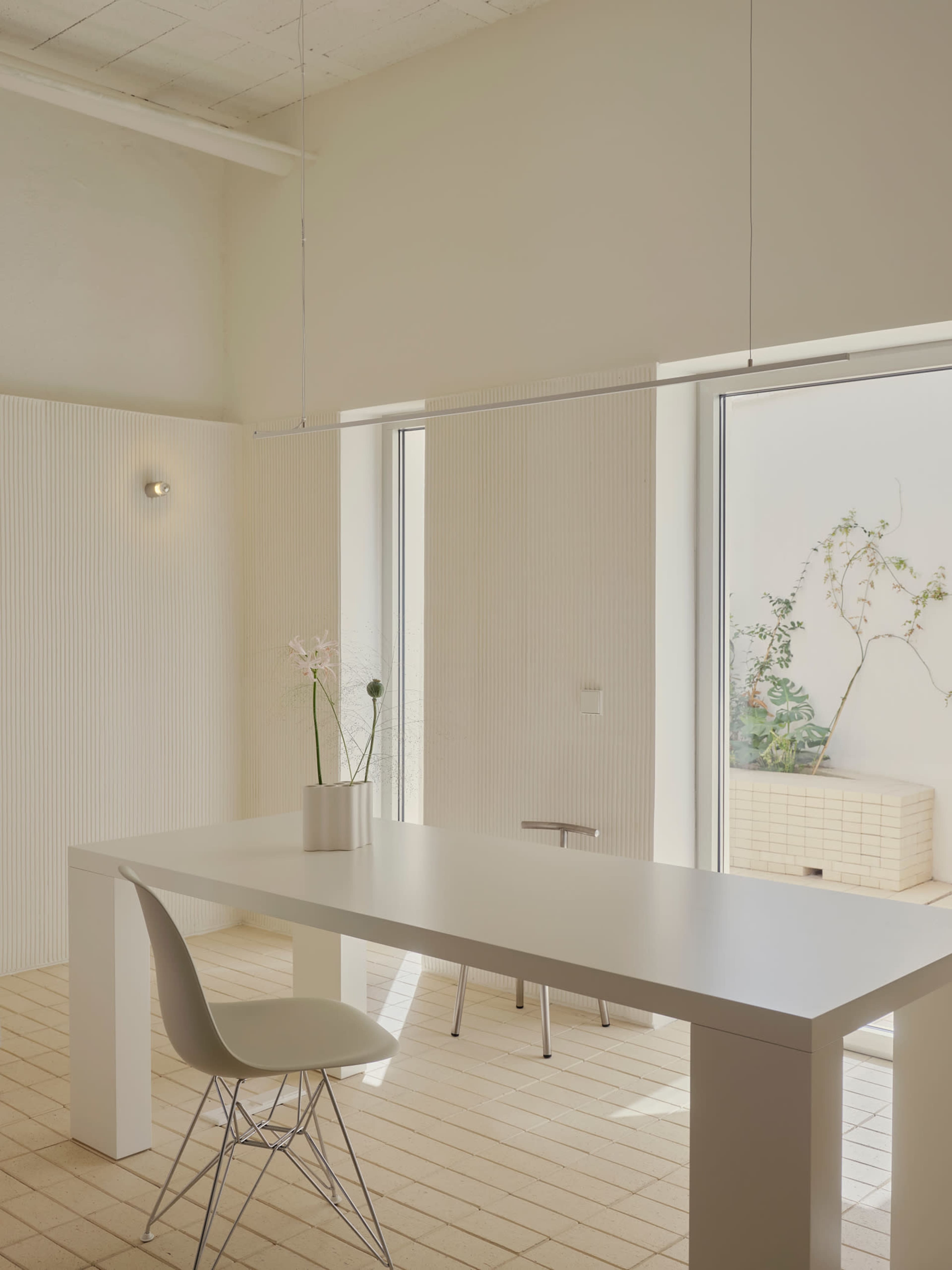 A minimalistic white table with a single chair is positioned in a bright room with large windows that overlook a small outdoor space.