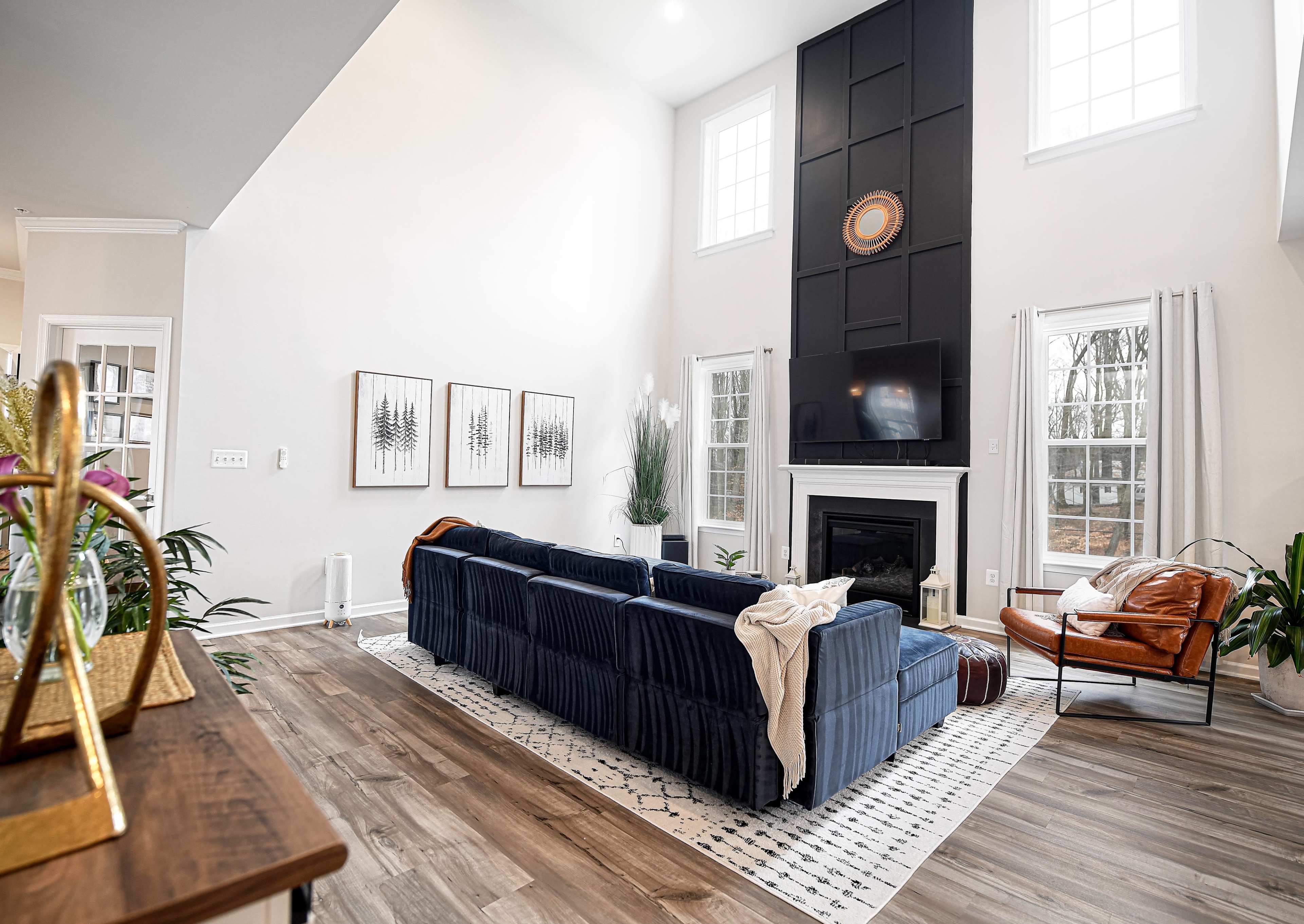 The image shows a bright living room with a tall ceiling, featuring a blue velvet sofa, a large wall-mounted TV above a fireplace, and large windows allowing natural light to enter.