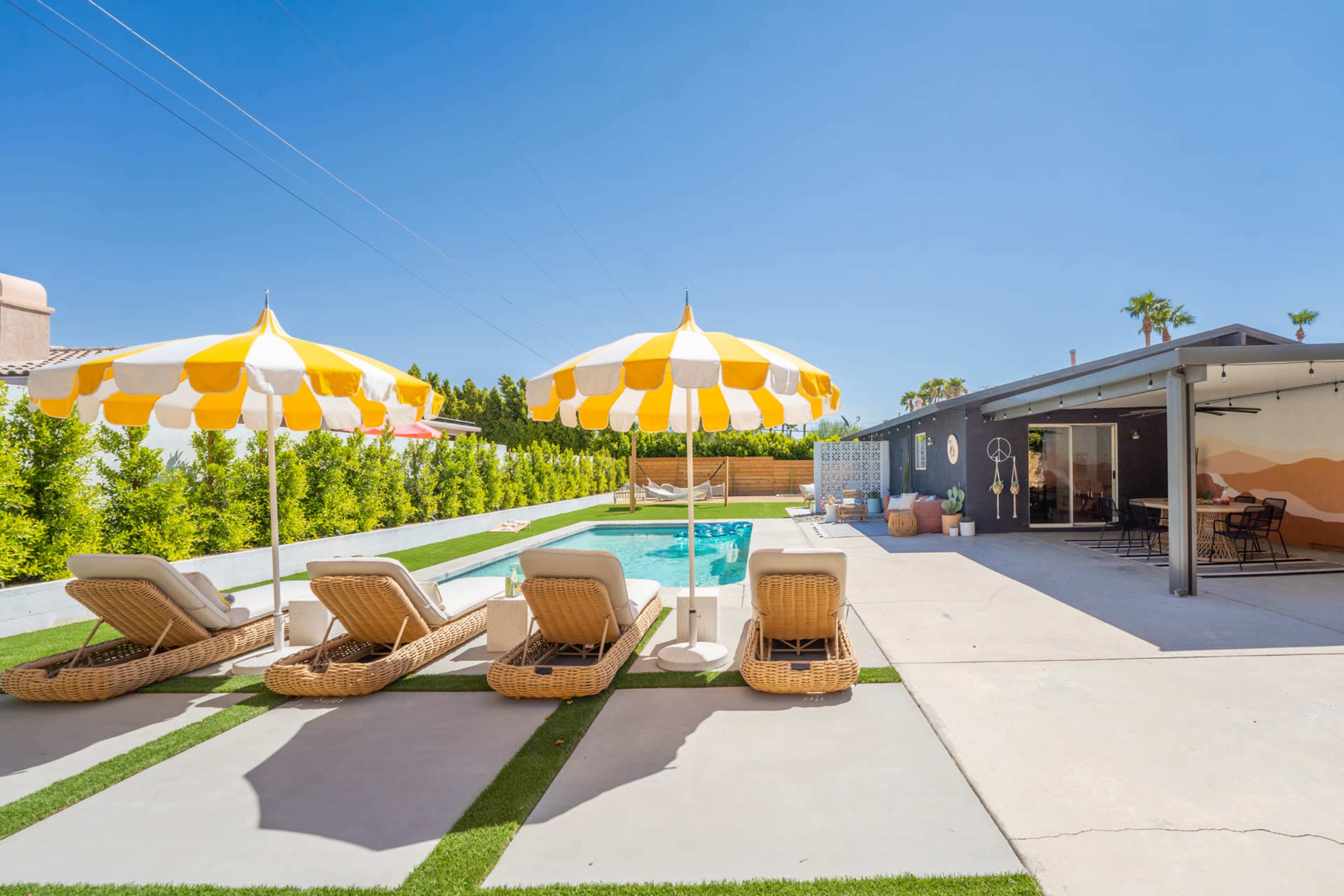 The image shows a pool area featuring lounge chairs with yellow and white striped umbrellas beside a swimming pool, surrounded by greenery and a modern home.