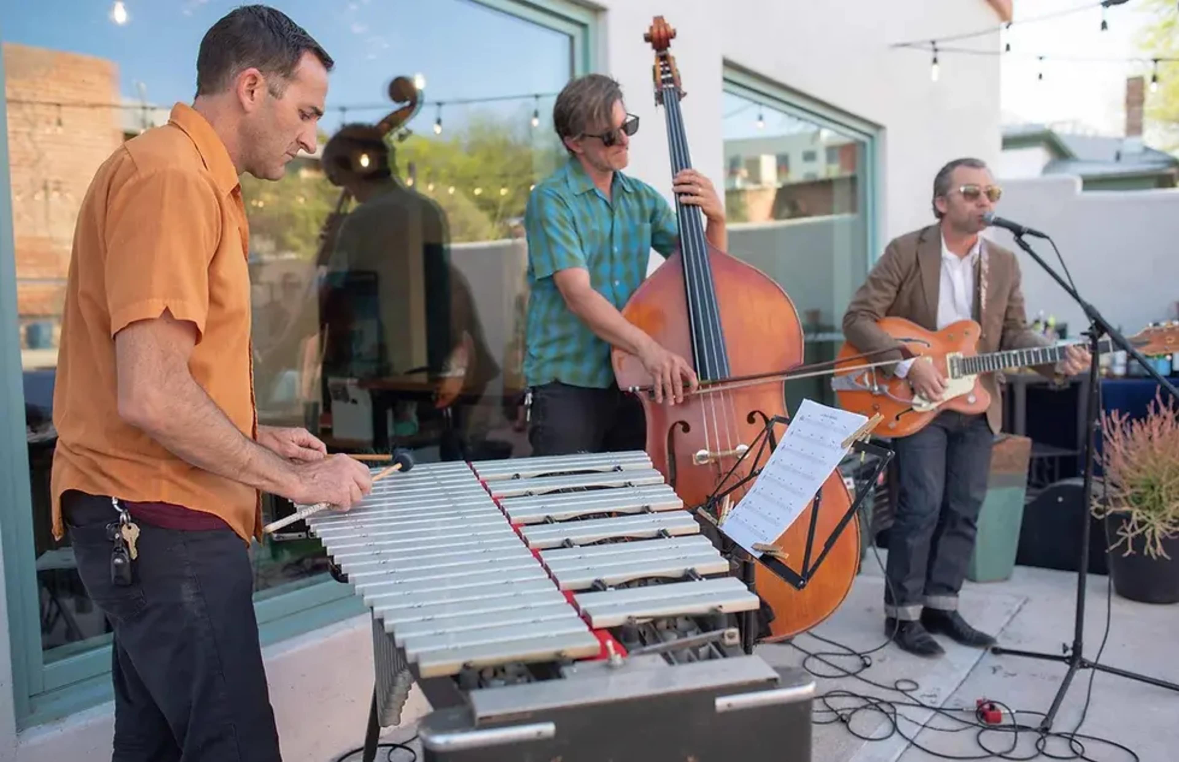 Three musicians perform at an outdoor venue, featuring a vibraphone, a double bass, and an electric guitar.