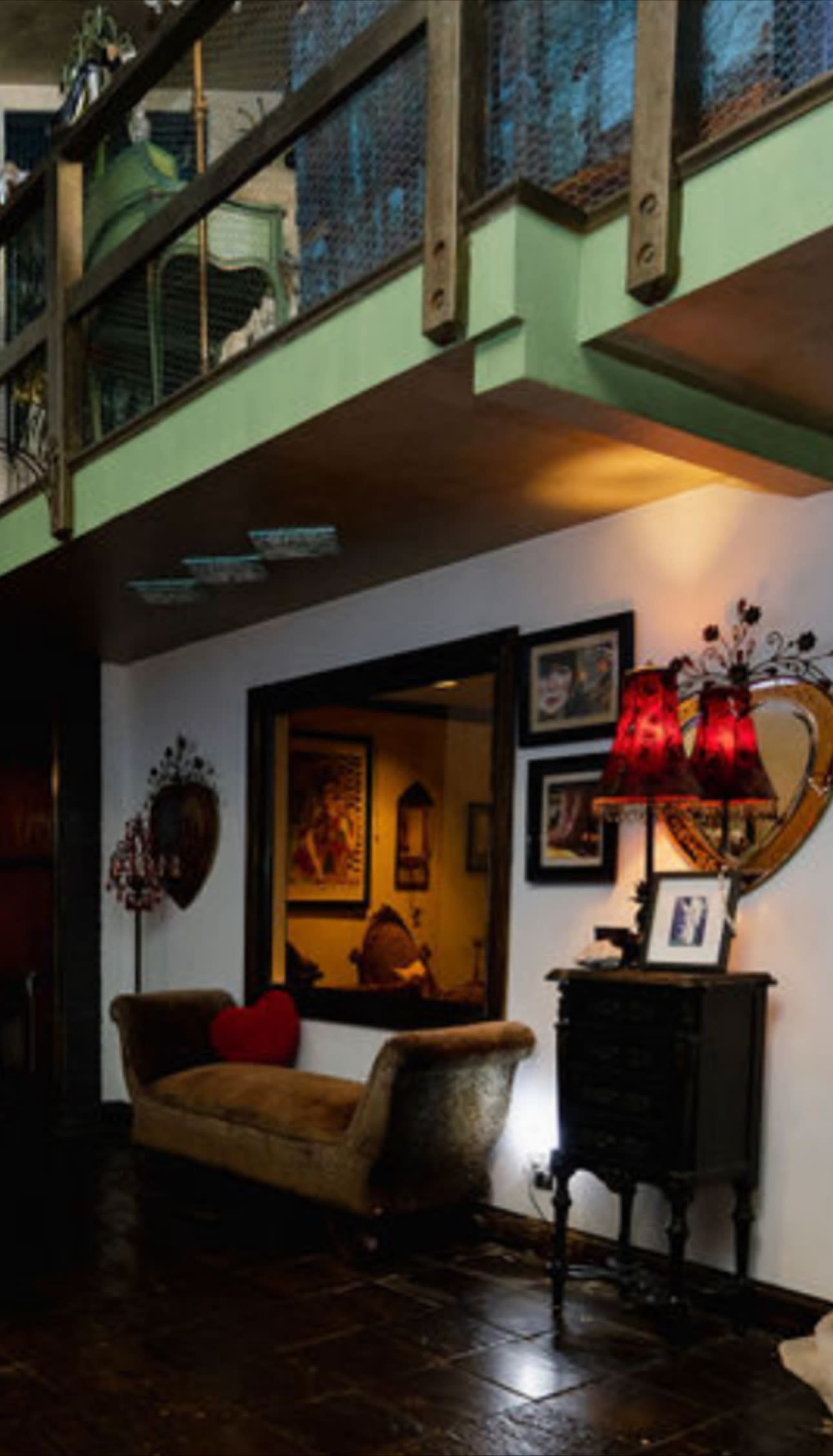 The image shows a cozy interior space featuring a brown sofa, a decorative red lamp, and a mirror reflecting artwork on the wall, with a balcony overhead.