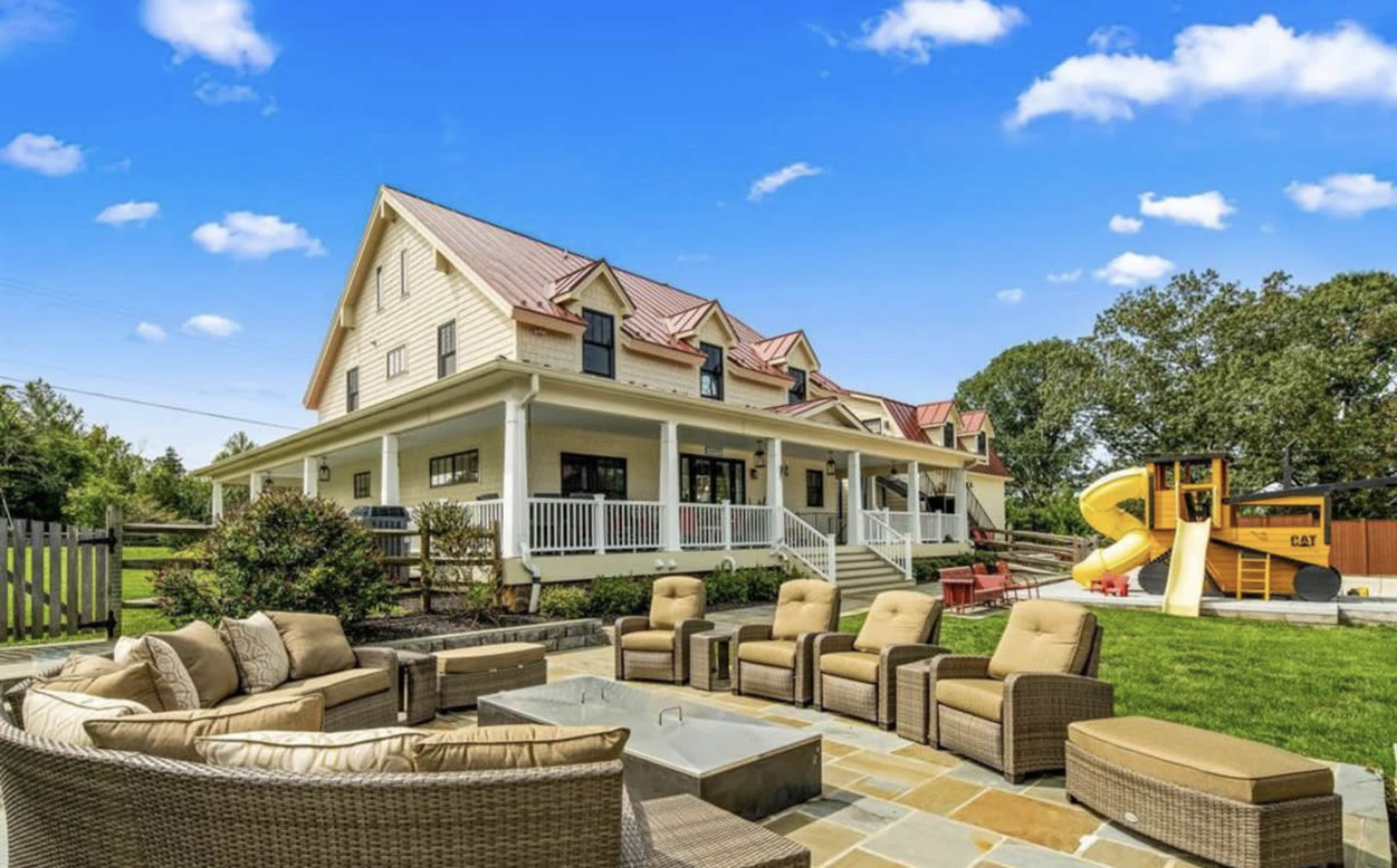 A spacious backyard features a seating area with wicker furniture surrounding a stone fire pit, alongside a large house with a red metal roof and a play structure.
