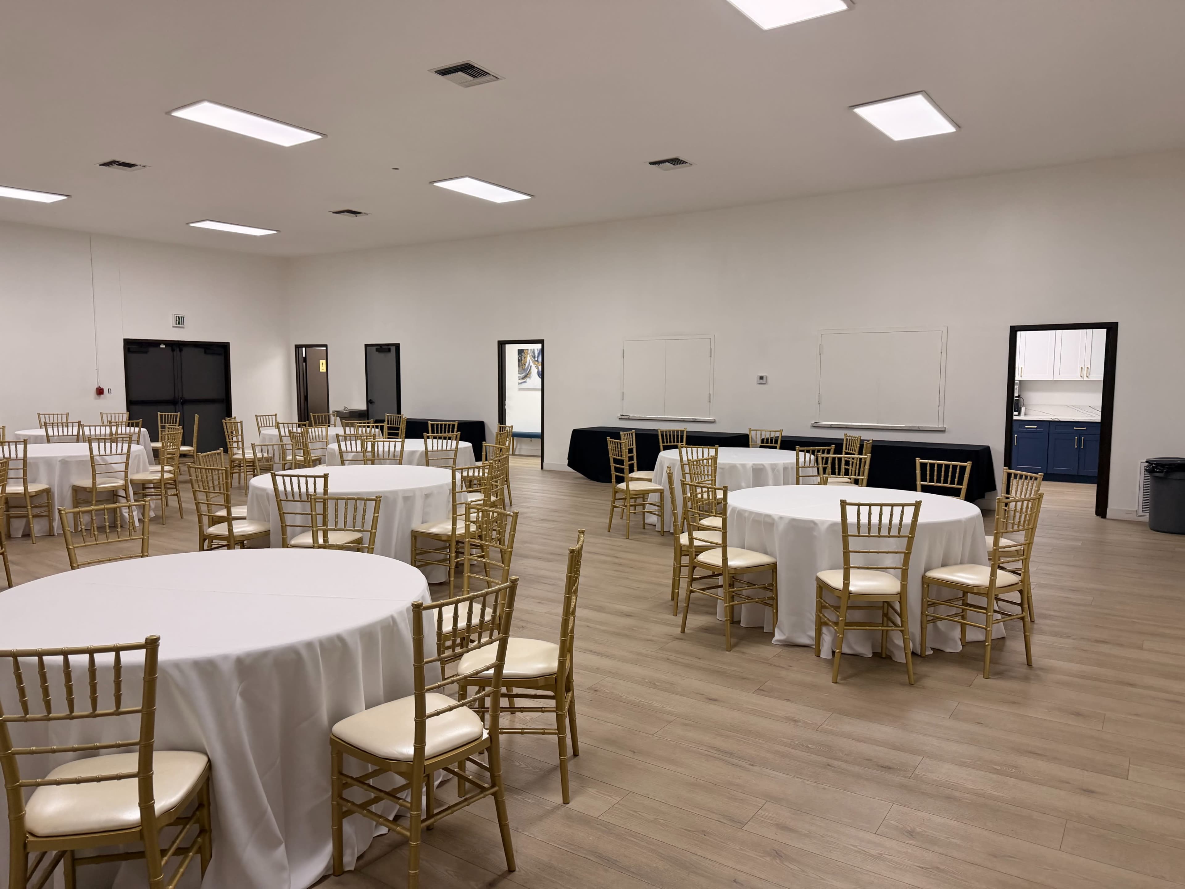 The image shows a spacious event room with multiple round tables dressed in white tablecloths and gold chairs arranged throughout the area.