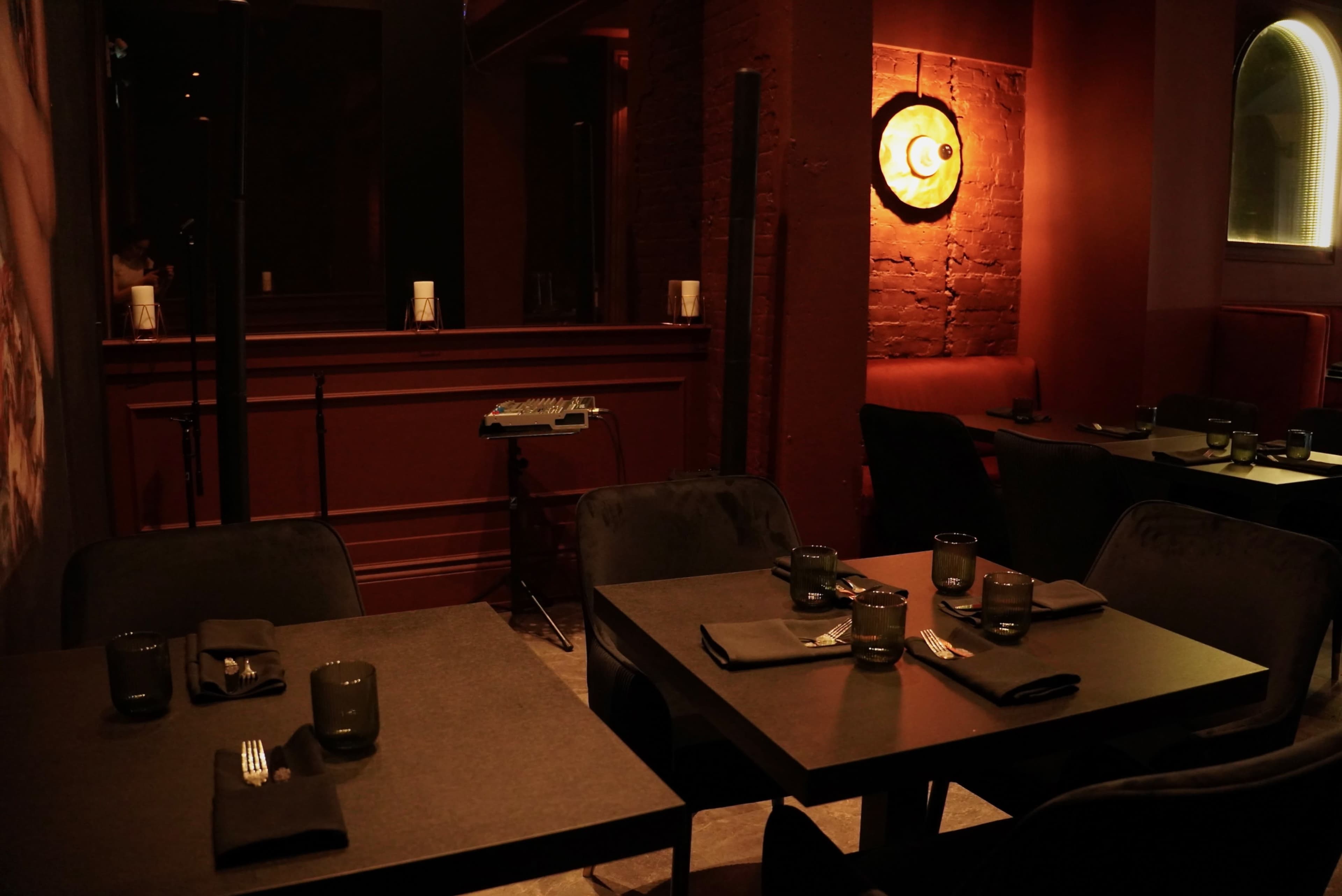 The scene shows a dimly lit restaurant with dark wood accents, tables set with glassware and utensils, and a warm orange wall light providing illumination.
