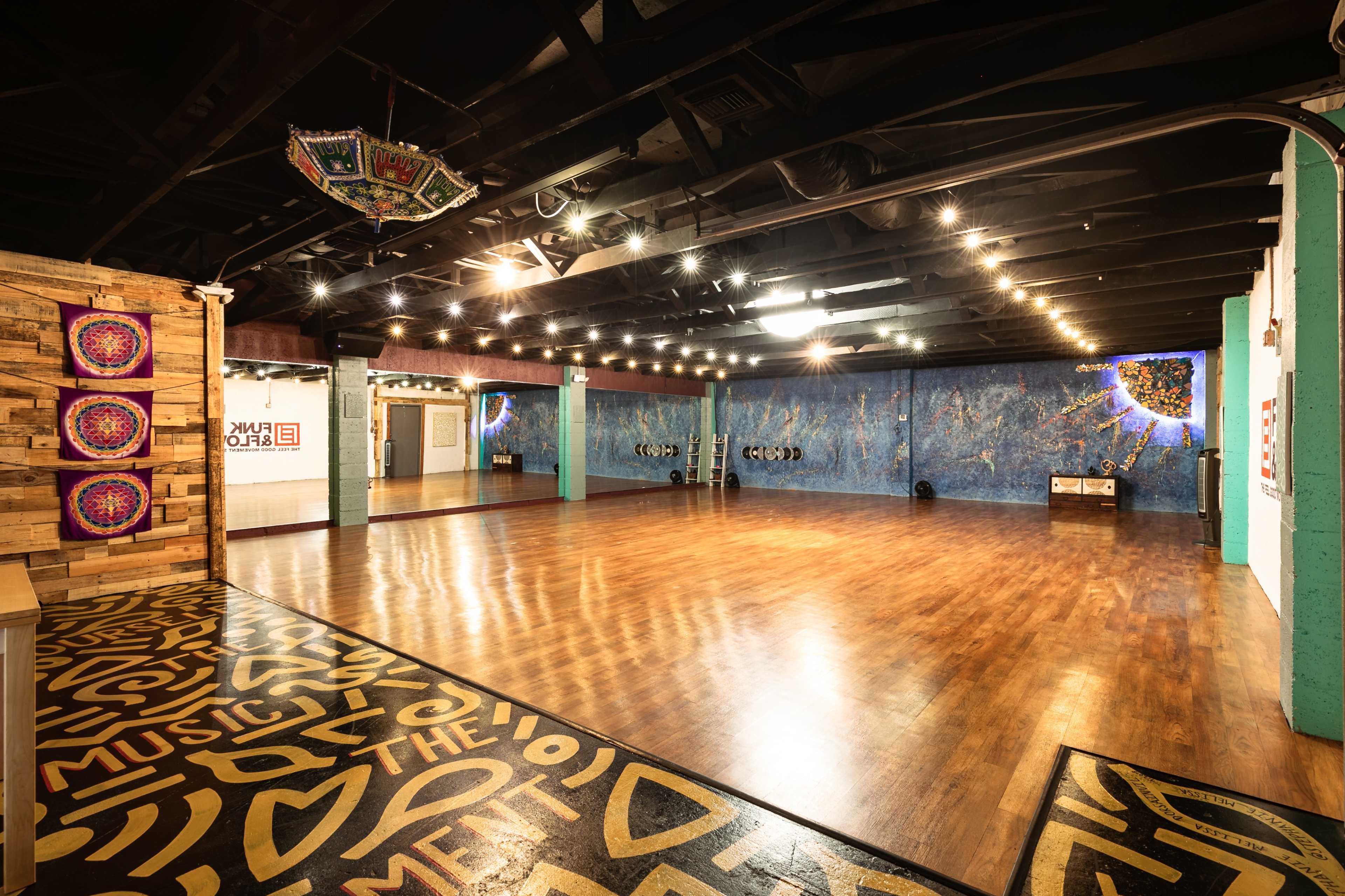 An empty dance studio features polished wooden floors, colorful wall murals, and dim overhead lighting.