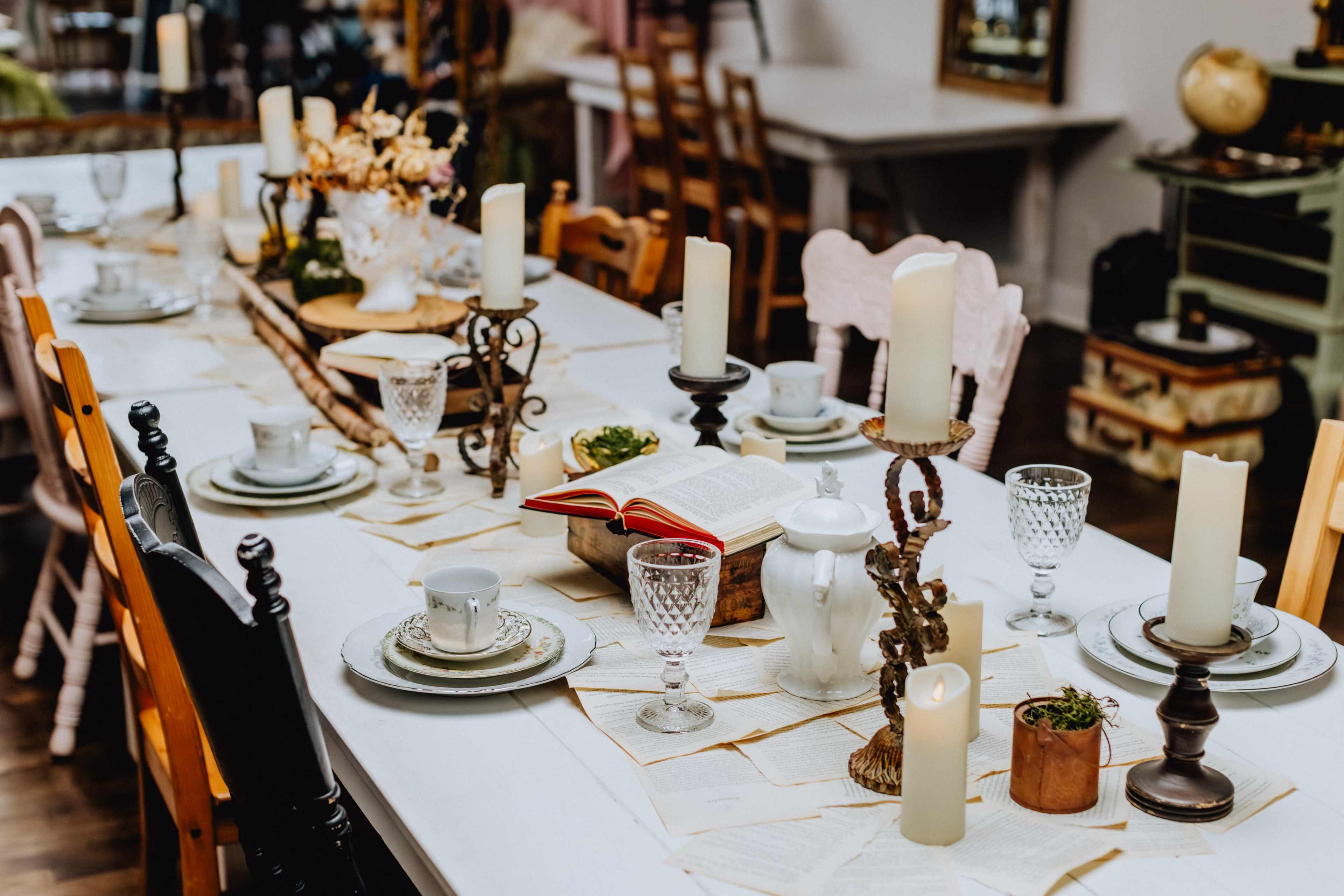 A long dining table is set with candles, decorative dishware, and an open book, surrounded by chairs in a cozy indoor setting.
