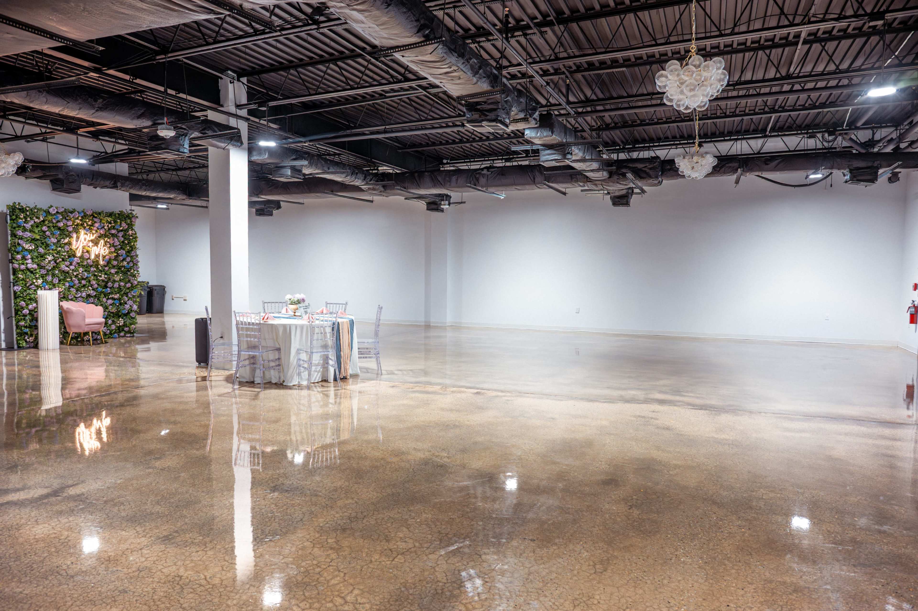 The image shows a spacious, empty indoor venue with polished concrete flooring, a few decorative elements, and a set of tables and chairs arranged in the center.