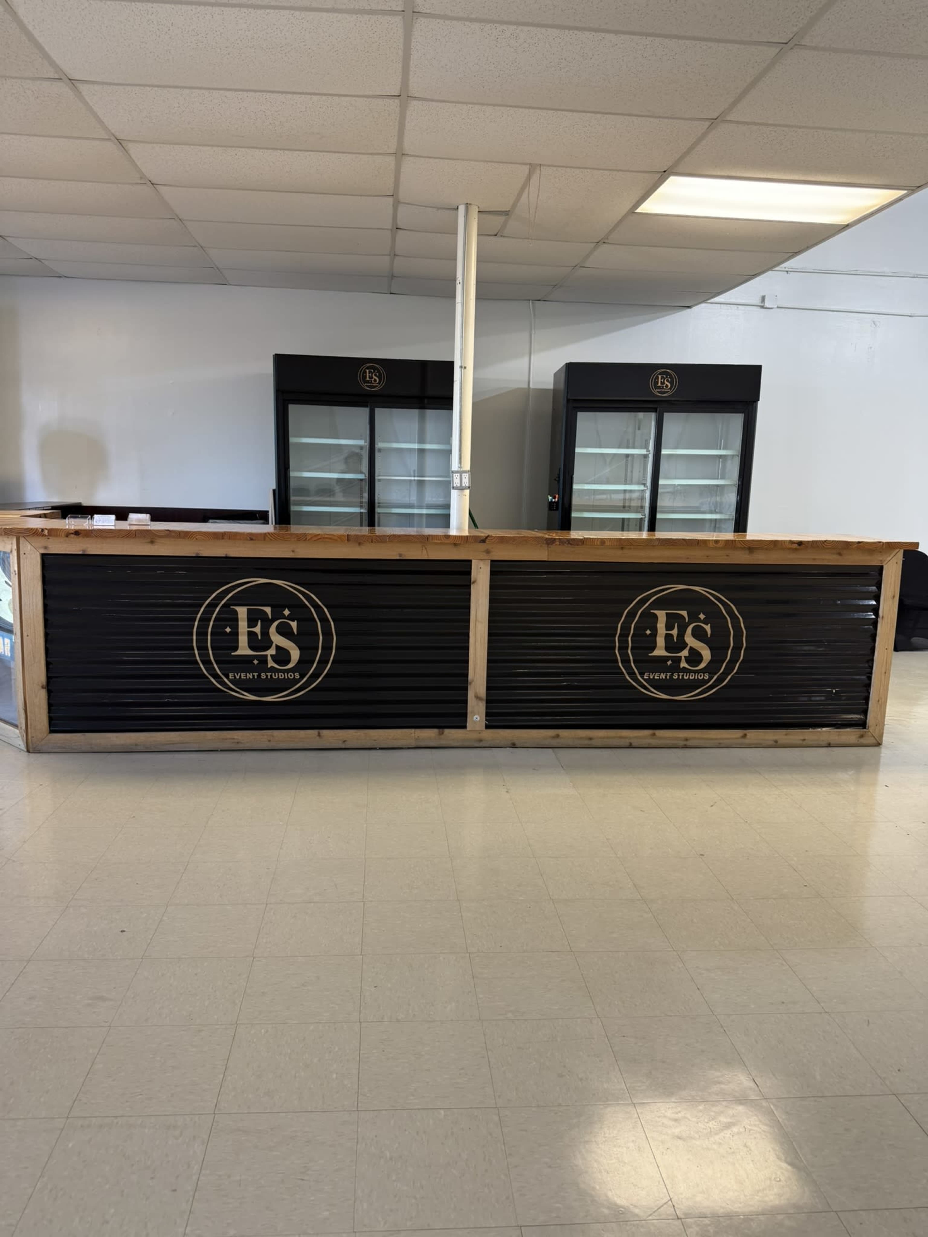 The image shows a wooden service counter with a black front displaying a circular logo, positioned in a room that has two glass-front refrigeration units behind it.