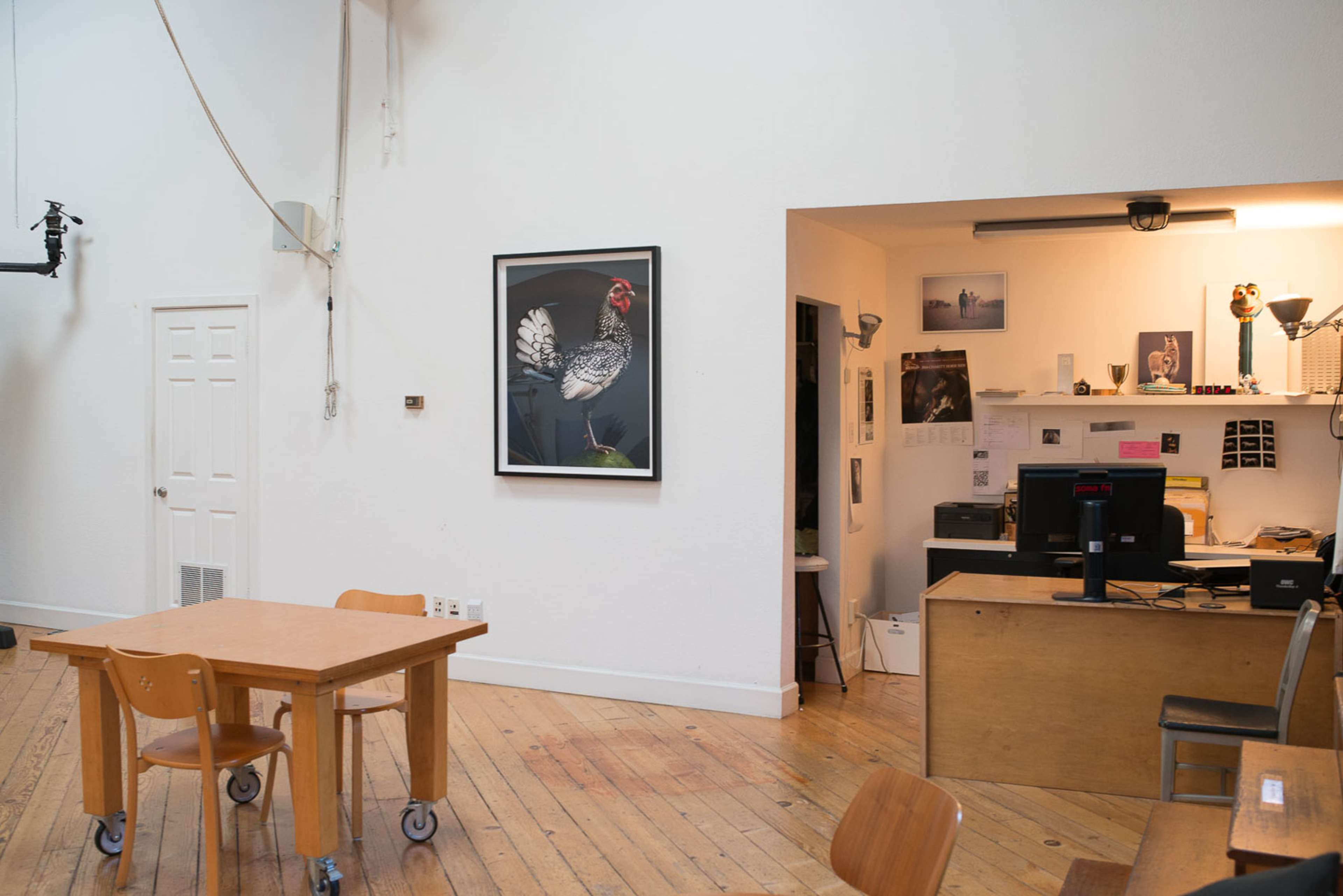 The image shows a bright, minimalist room with wooden floors, featuring a large framed picture of a chicken on the wall and a desk with a computer in the corner.