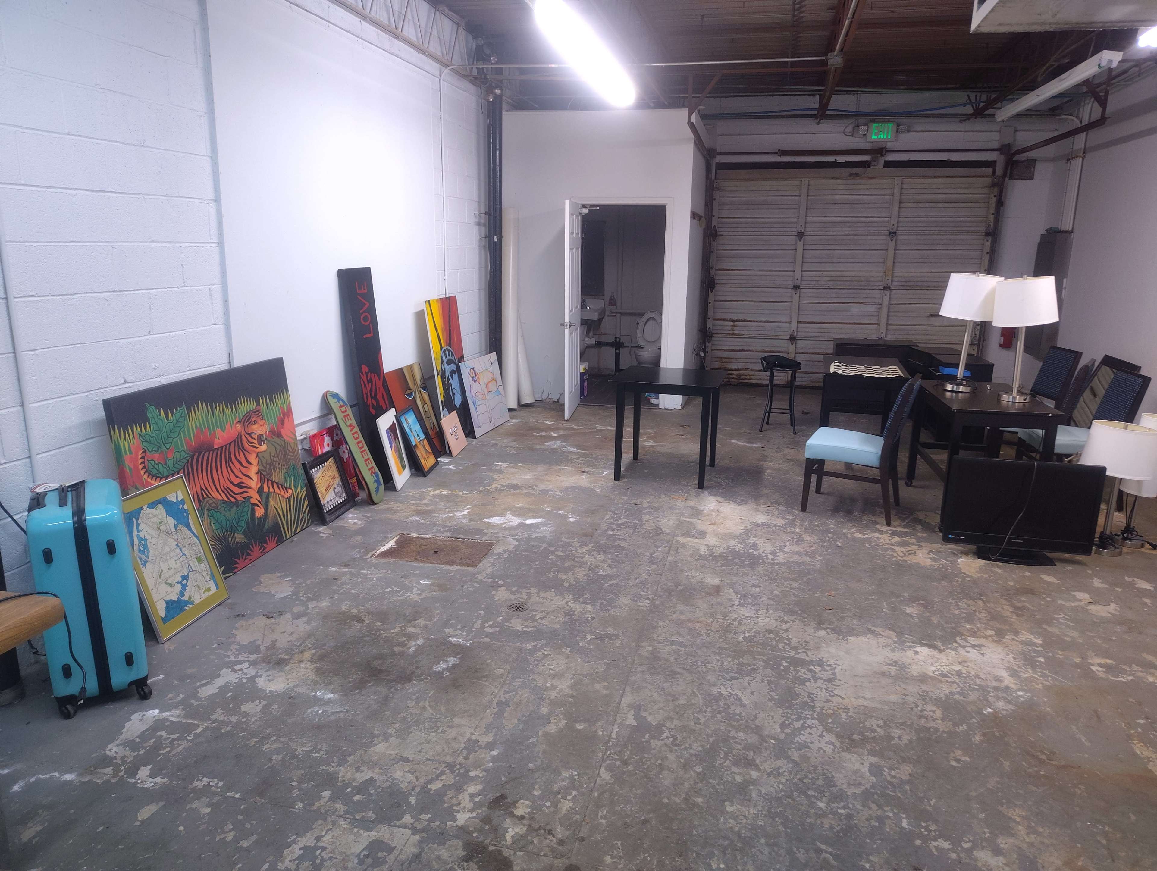 The image shows an unfinished room with a concrete floor, featuring several framed artworks leaning against the wall, a table and chairs, and two lamps in a separate area.