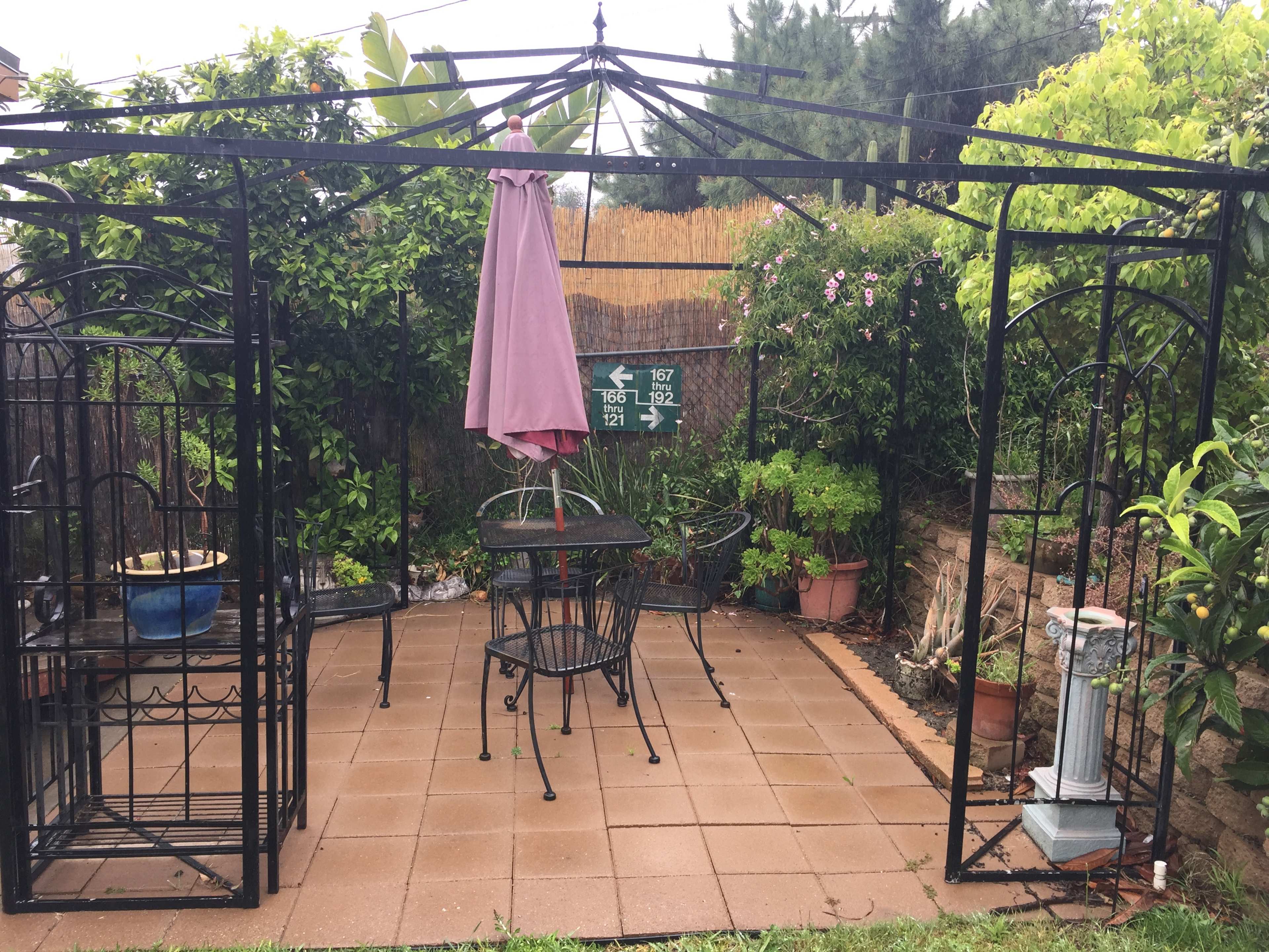 A patio area features a black metal frame with a pink umbrella, surrounded by potted plants and two black chairs and a table on tiled flooring.
