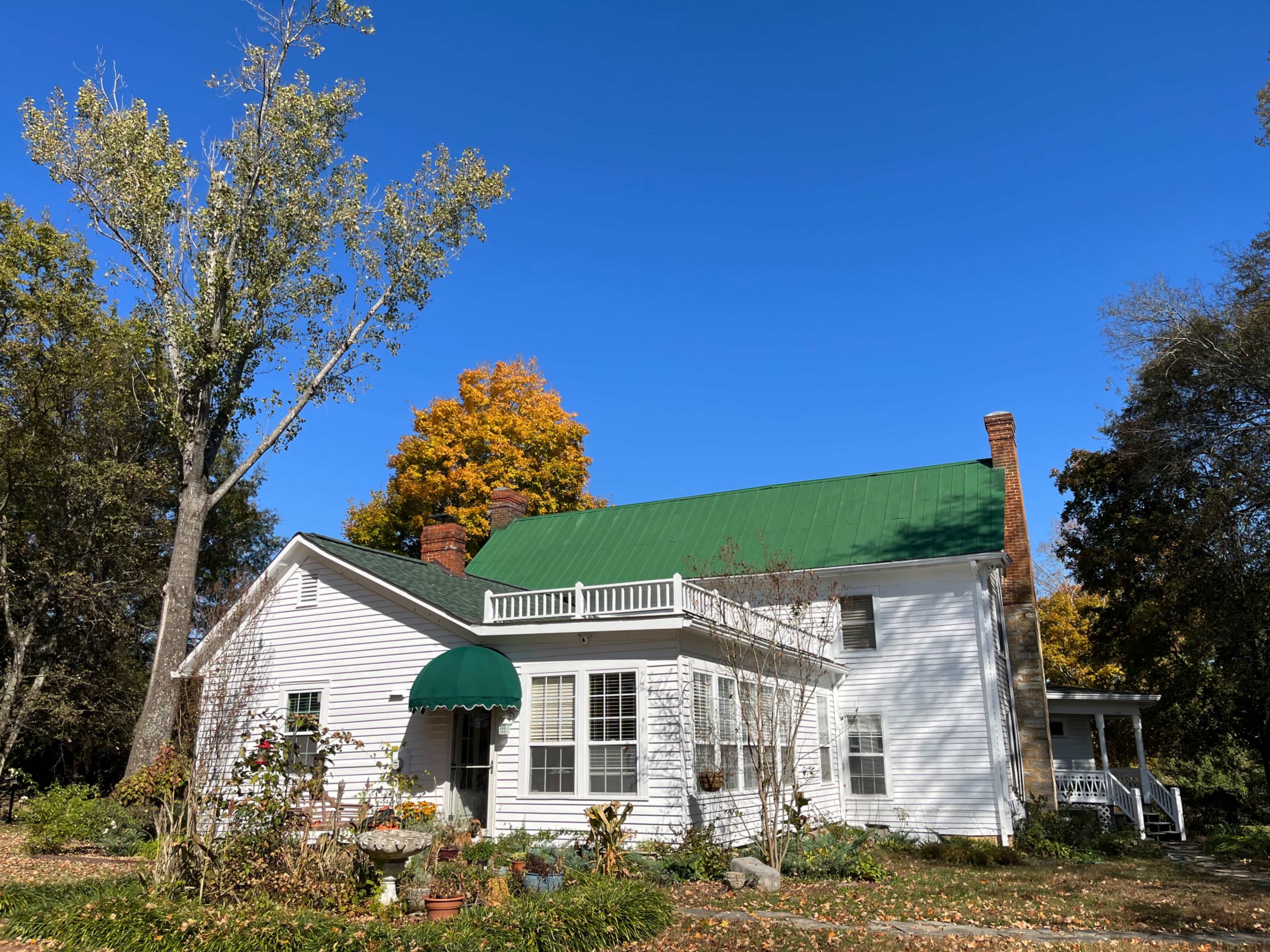 A white house with a green metal roof and a porch is surrounded by trees, some of which have autumn leaves.