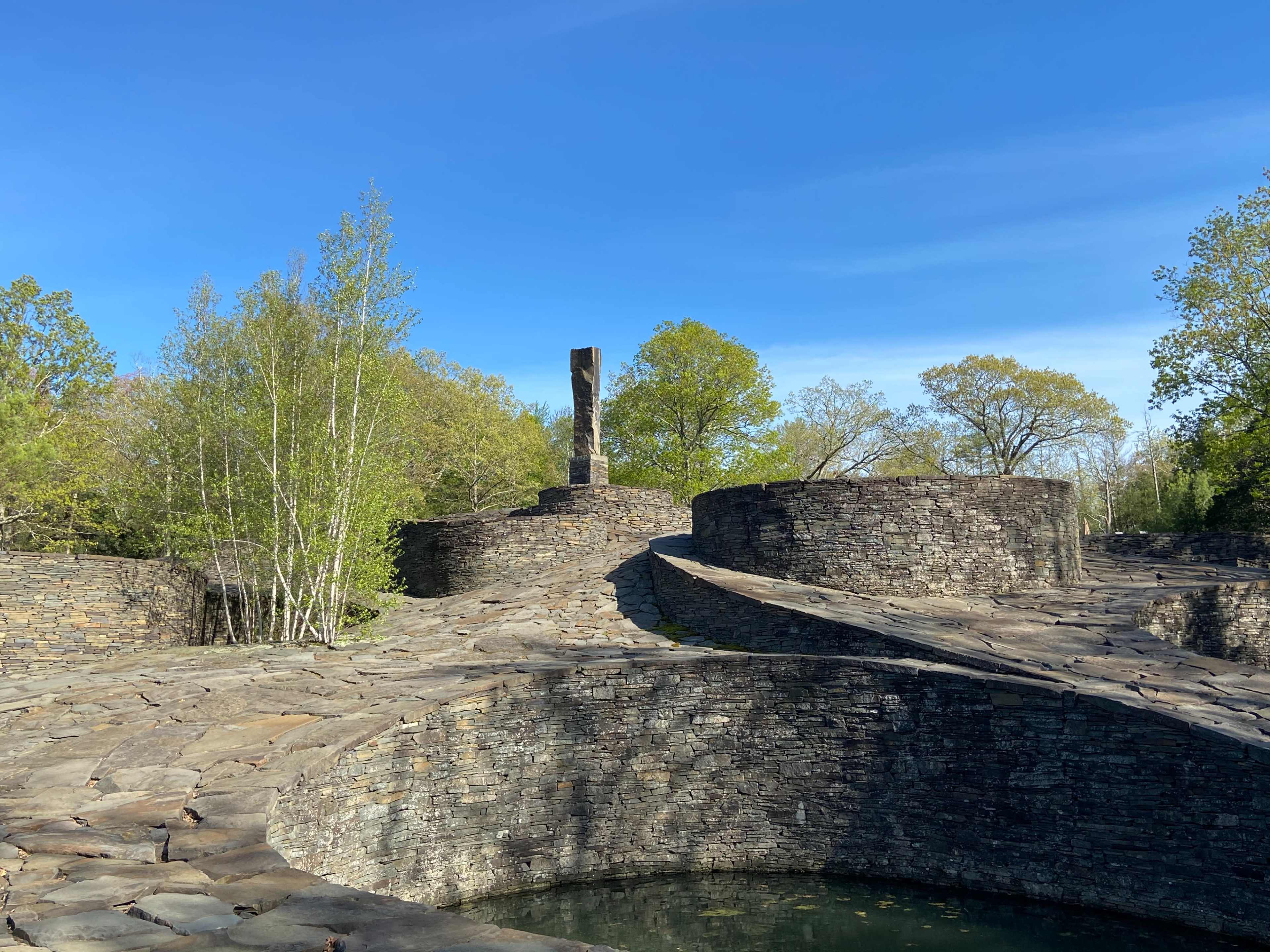 A stone landscape featuring circular formations, a central vertical pillar, and surrounding greenery under a clear blue sky.
