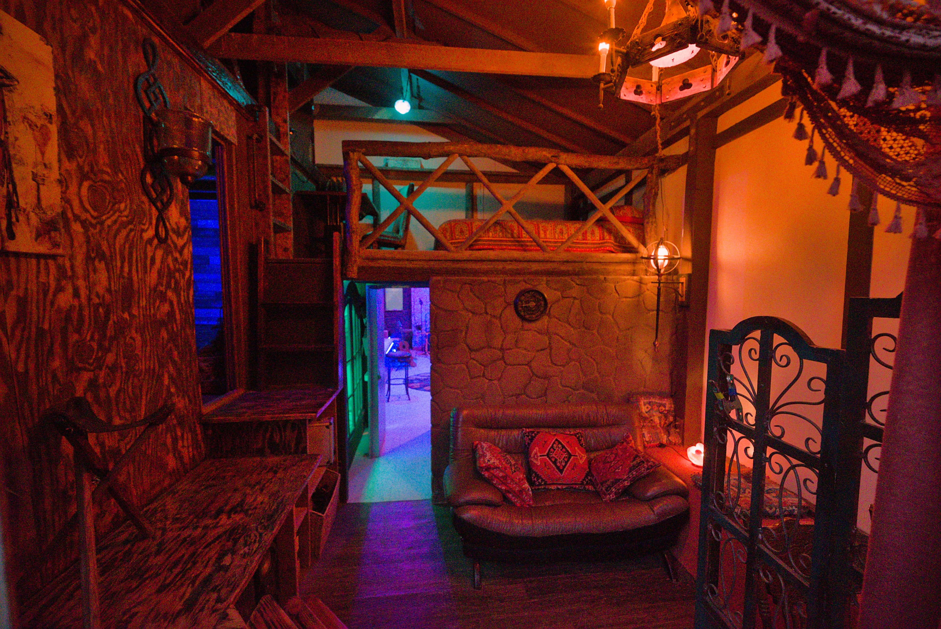 The image depicts a cozy, rustic room featuring a sofa, wooden decor, and a loft area under a sloped ceiling with colorful lighting.