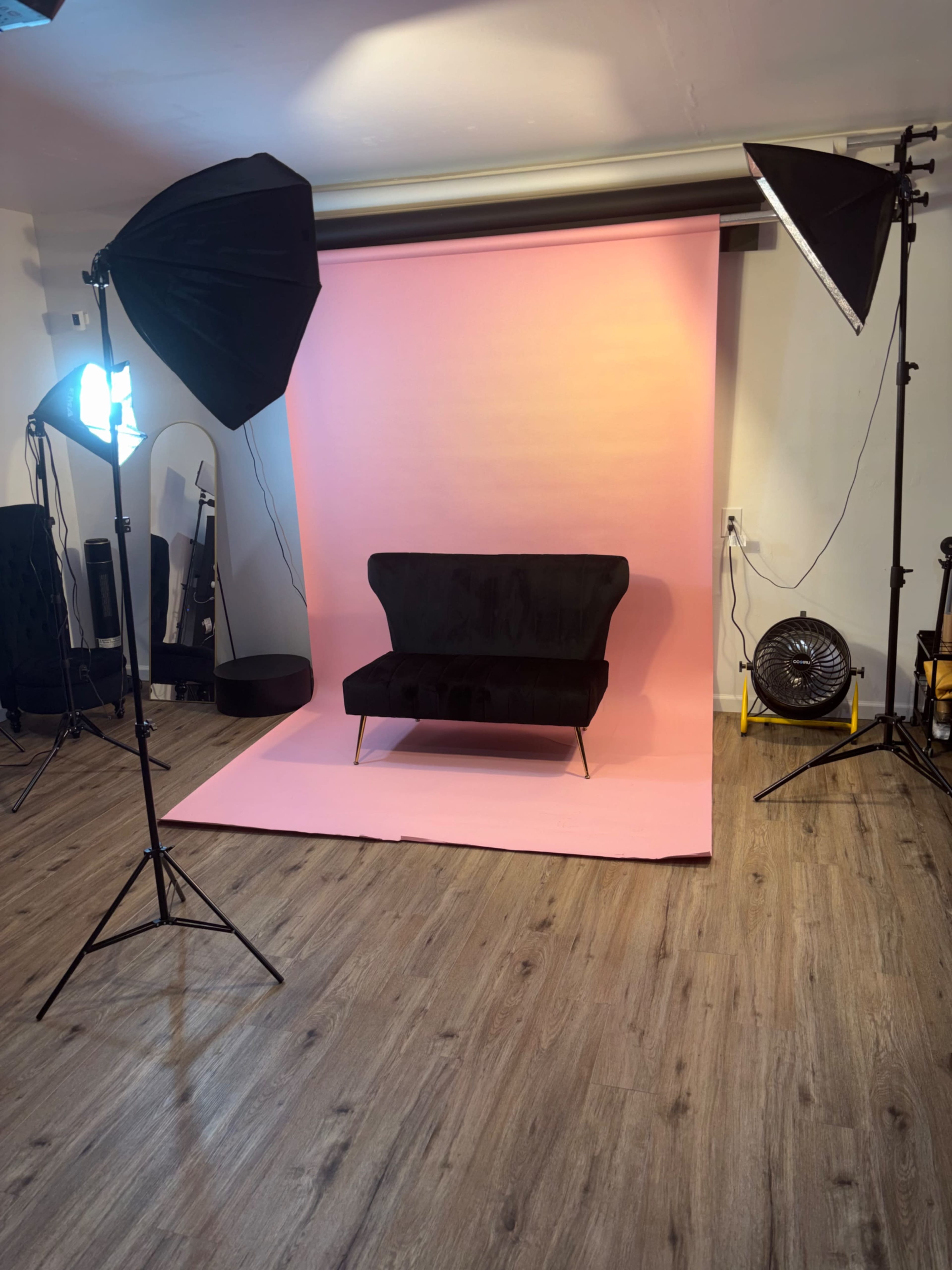 The image shows a photography studio setup featuring a black sofa on a pink backdrop, with two softbox lights and a fan positioned nearby.
