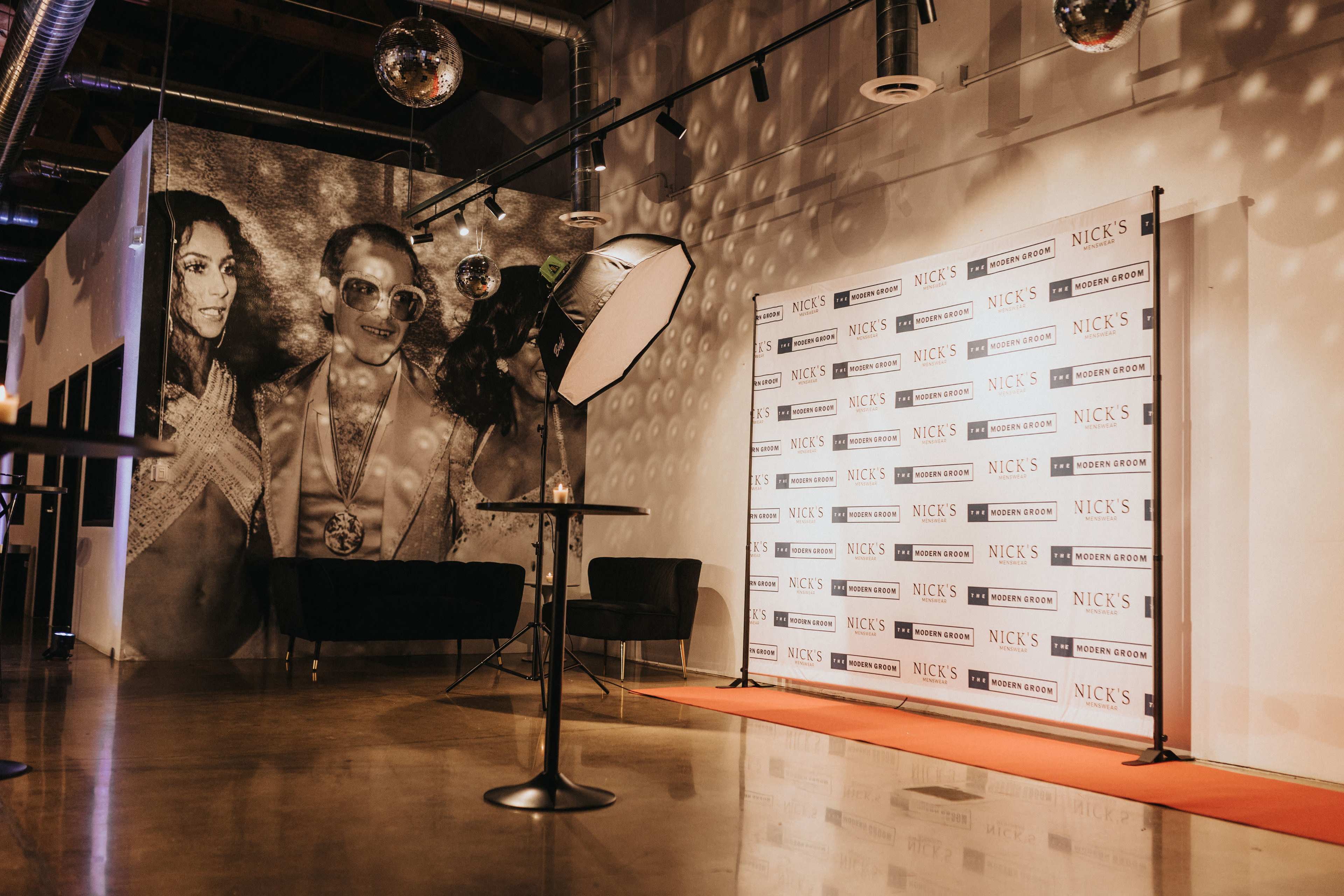 The scene features a photo backdrop with a red carpet, a spotlight, and a large mural of three individuals on the wall in a well-lit venue.