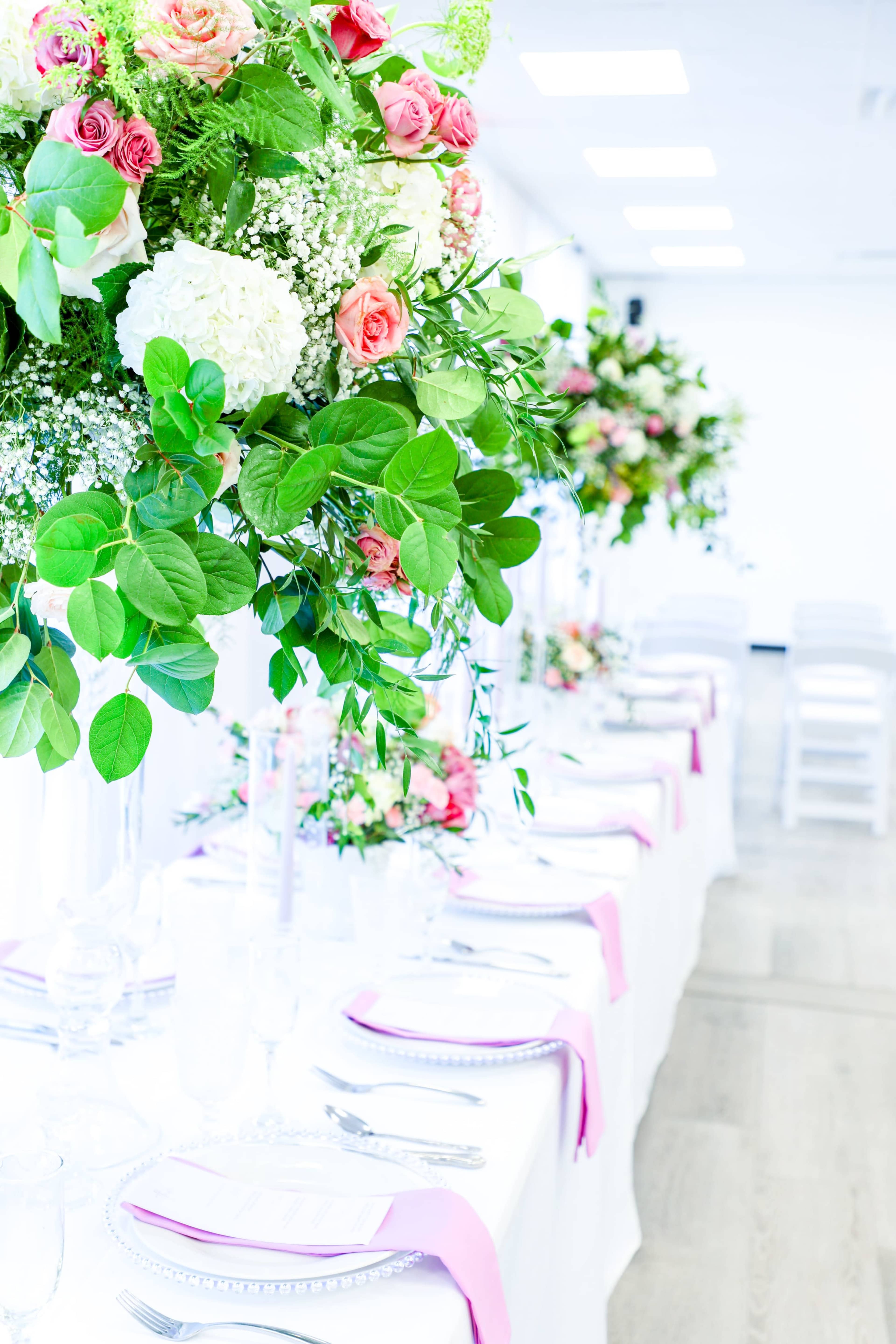 A long banquet table is set with white tablecloths, pink napkins, and tall floral centerpieces featuring greenery and roses.