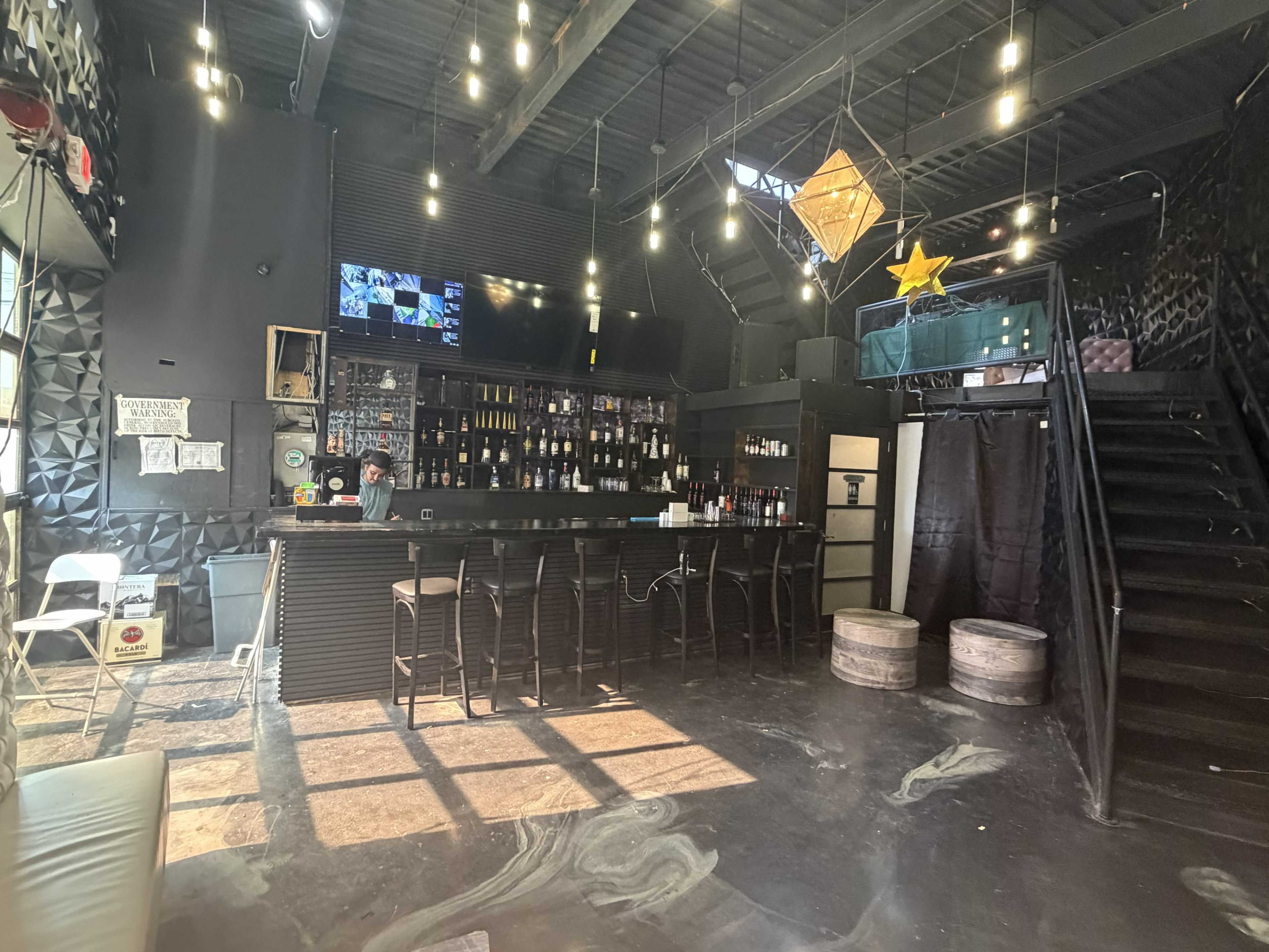 The image shows an empty bar area with a black countertop, shelves filled with bottles, and a staircase leading to a mezzanine level, illuminated by pendant lights.