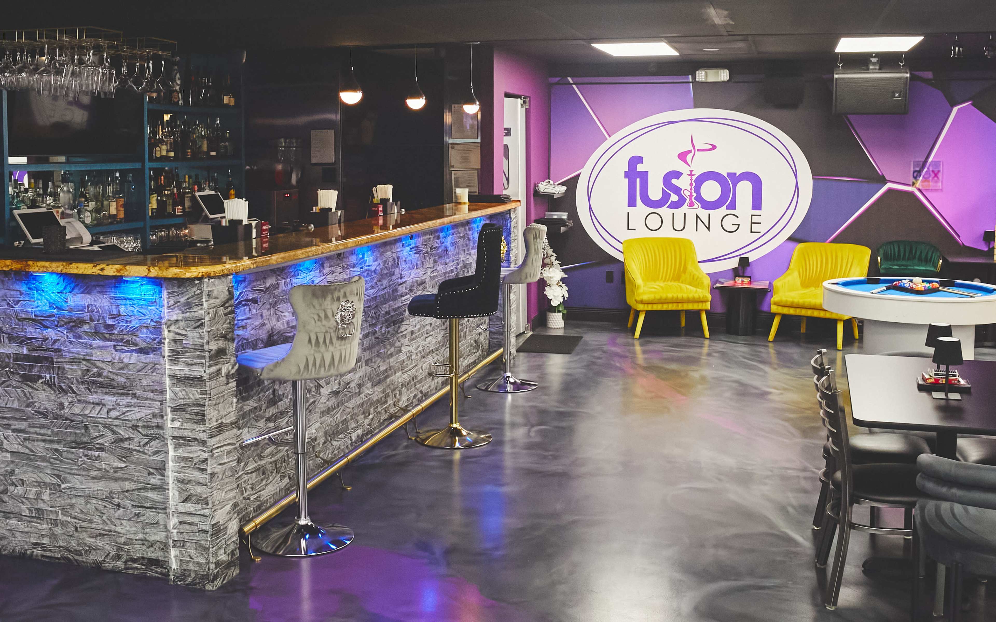The image shows a lounge interior featuring a bar with high stools, yellow seating, and a blue-lit area, all set against a purple wall.