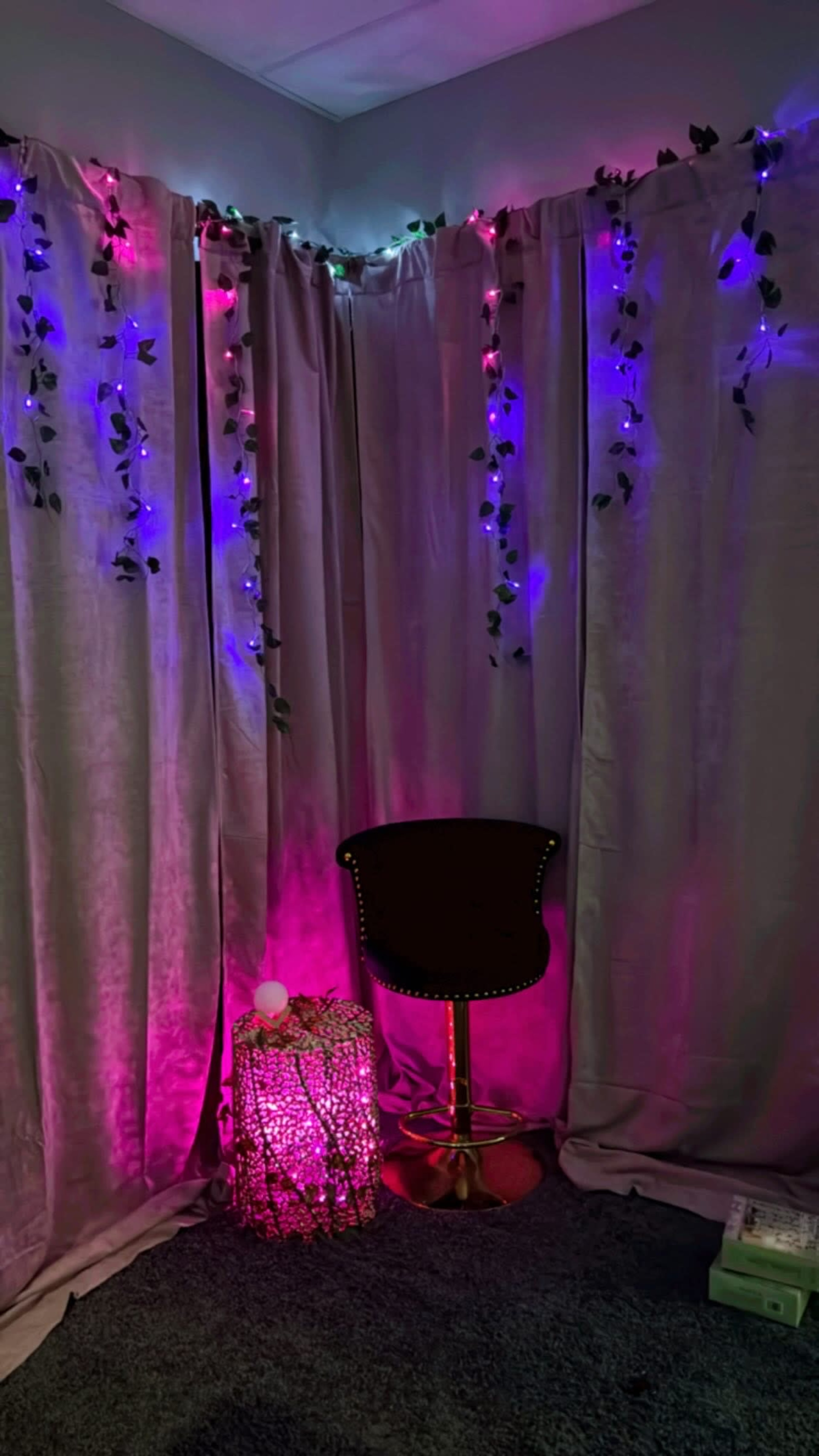 A corner decorated with colorful lights, a black stool, and a decorative basket, all set against a backdrop of curtain drapes.