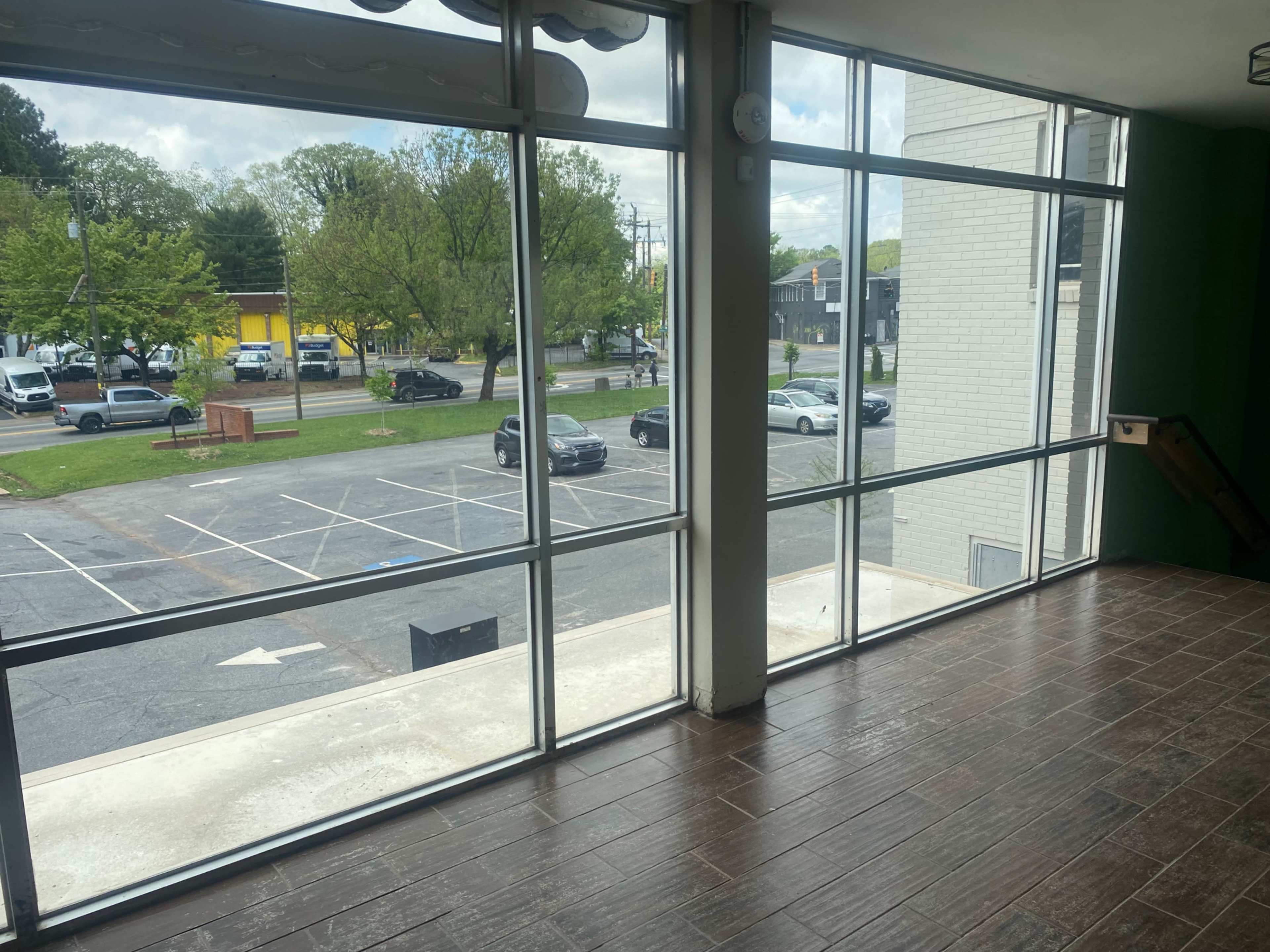 The image shows a spacious room with large windows overlooking a parking lot and a street with parked cars and trees.