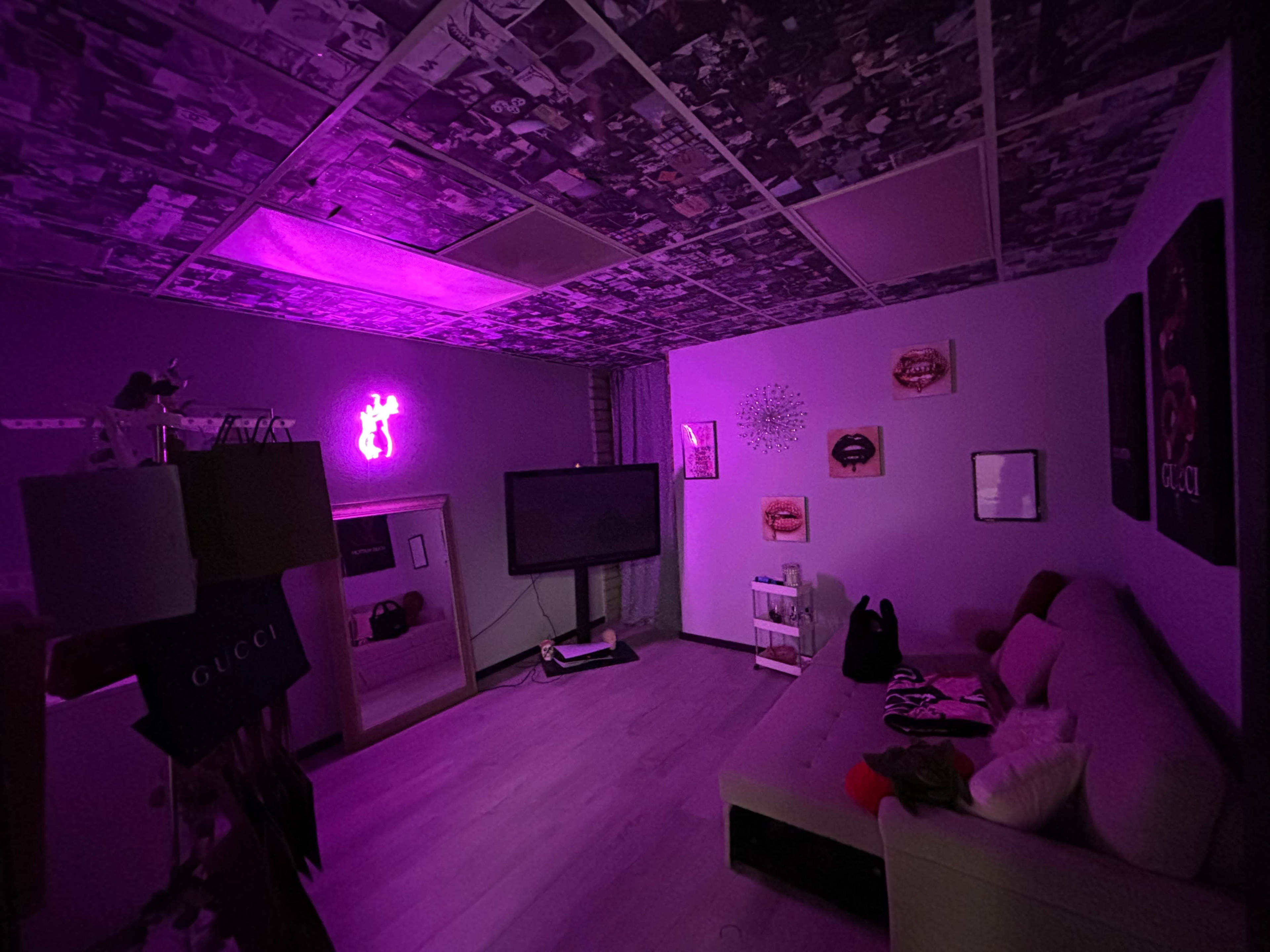 A dimly lit room features a couch, a television, and walls adorned with artwork, illuminated by a purple neon light.
