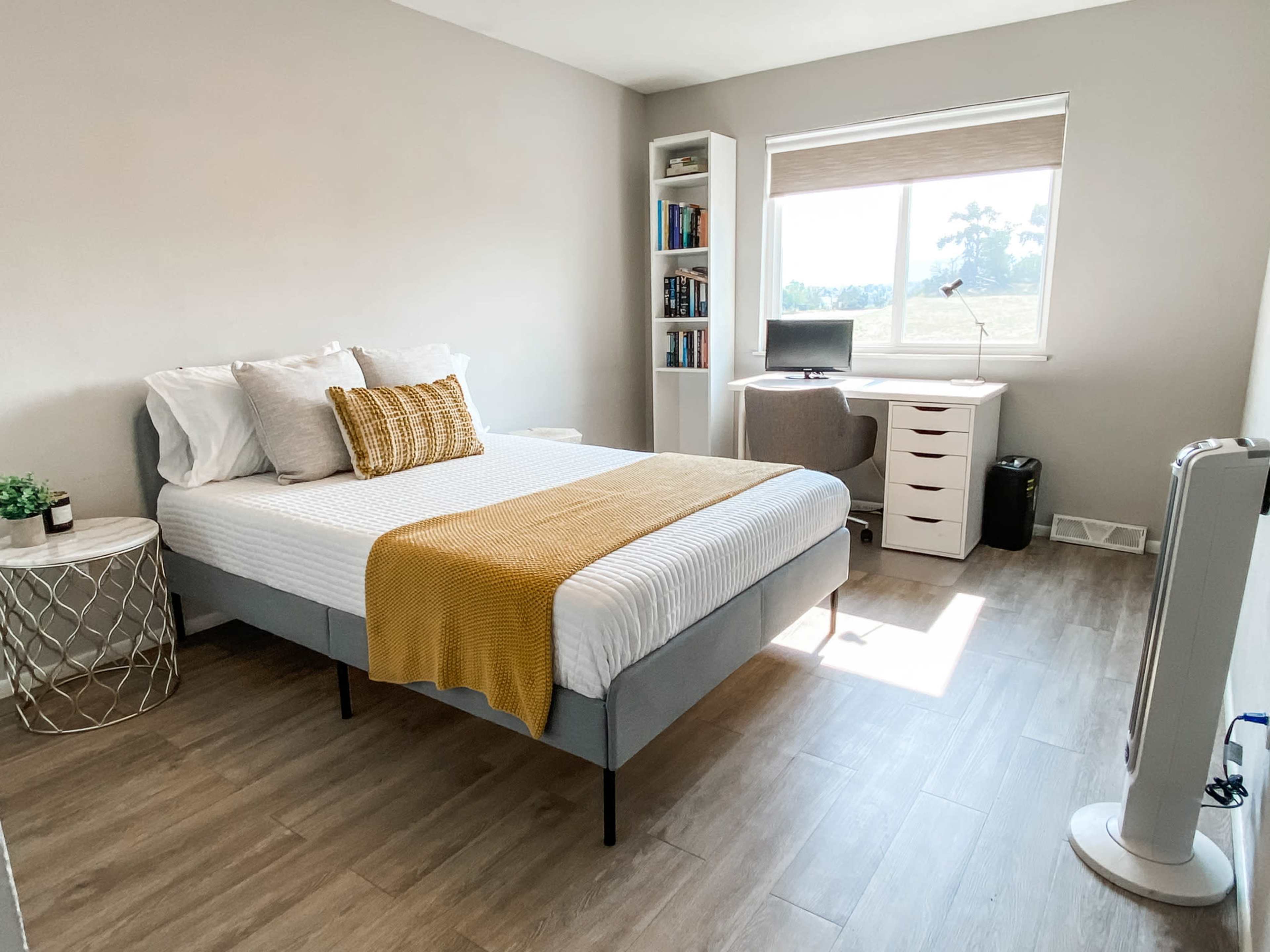A simple bedroom featuring a bed with a yellow throw, a small side table, a desk with a computer, and a bookshelf against a wall, illuminated by natural light from a nearby window.