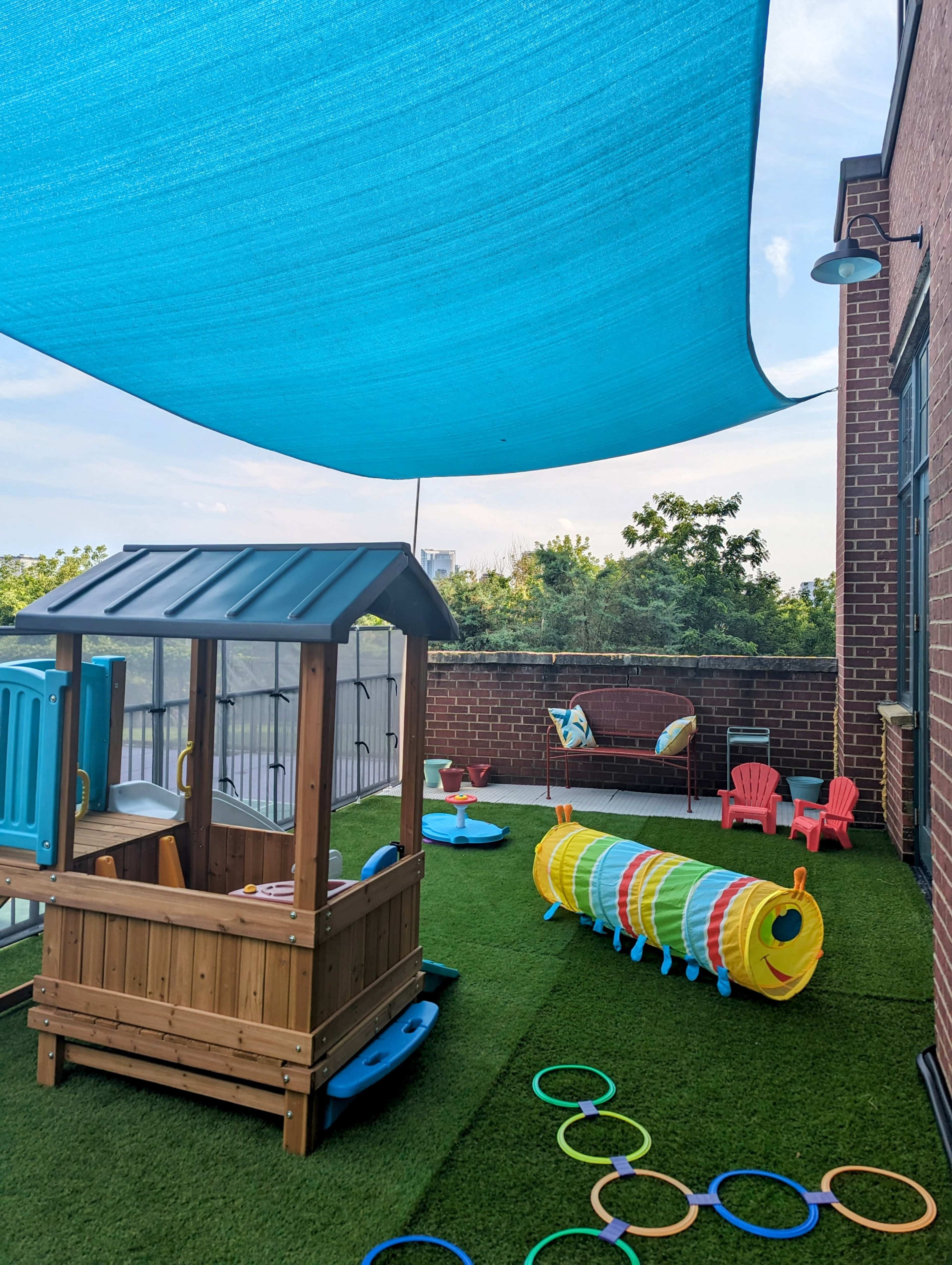 A covered play area features a colorful playhouse, a tunnel, and soft flooring, surrounded by greenery and a brick wall.
