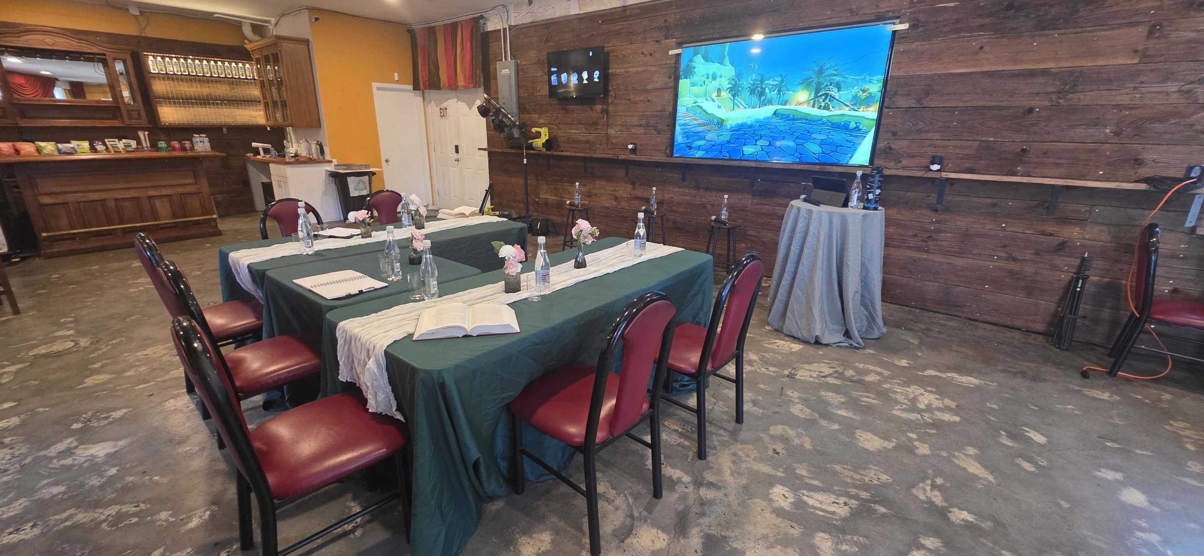 The image shows a room set up for a meeting or presentation, featuring two tables with green tablecloths, chairs, water bottles, and a large screen displaying a tropical scene on a wooden wall background.