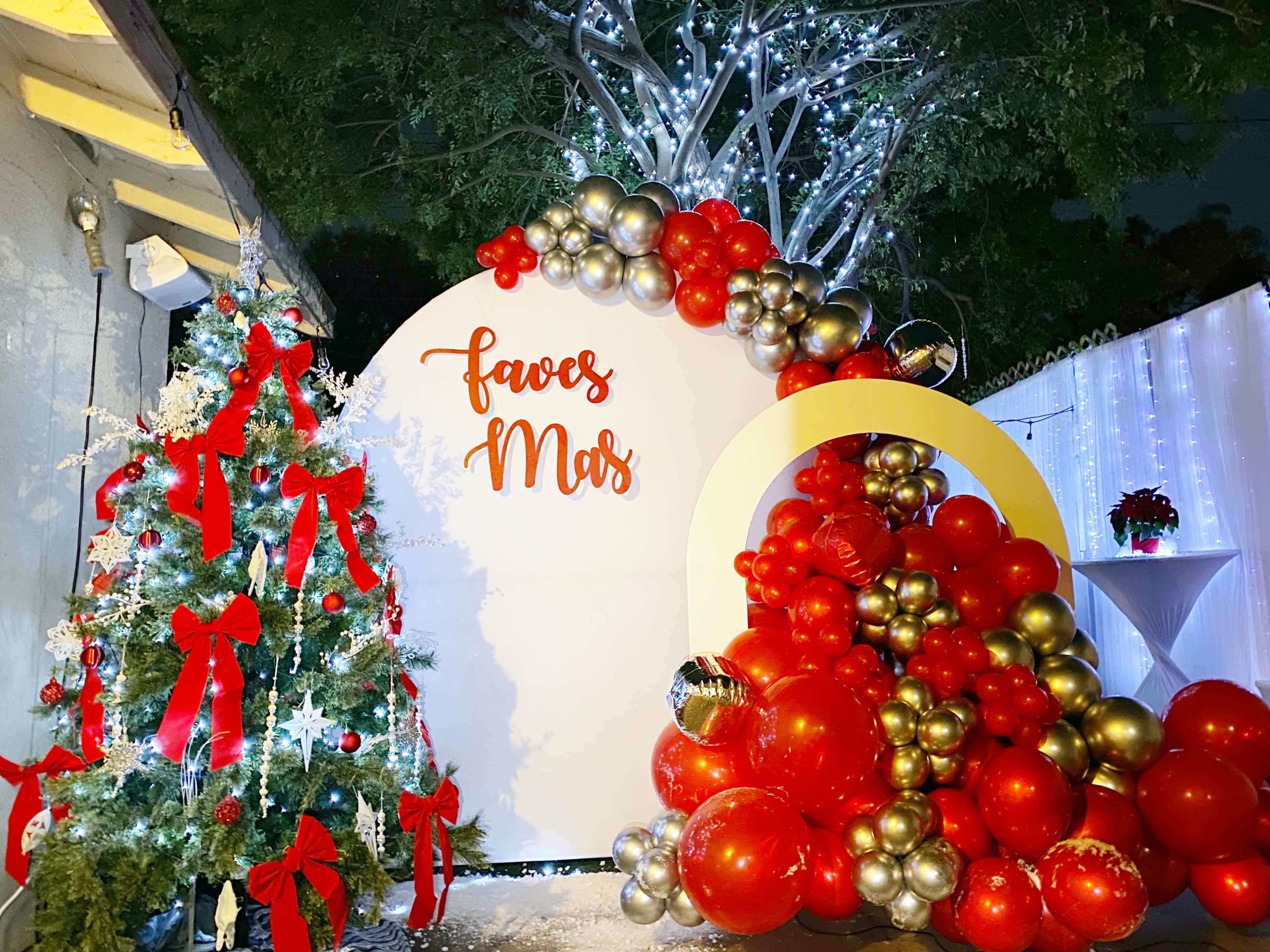 A festive display with a decorated Christmas tree on one side and a large balloon arrangement in red and gold on the other, set against a backdrop that says "faves Mar."